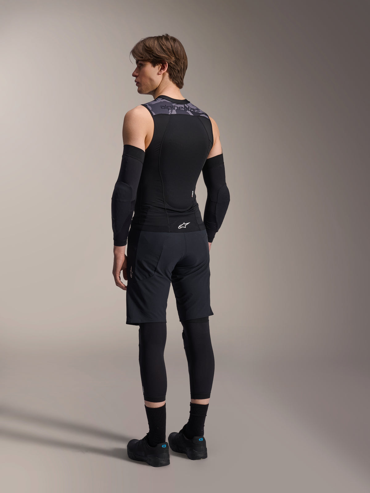 Mandlig model set bagfra iført Alpinestars A-Motion Plasma beskyttelsesvest, Black Camo, ærmeløst mesh-design med integreret rygbeskytter, gråt camouflage-mønster på skuldrene, kombineret med sorte albuebeskyttere og mountainbike-shorts, professionelt cykeludstyr