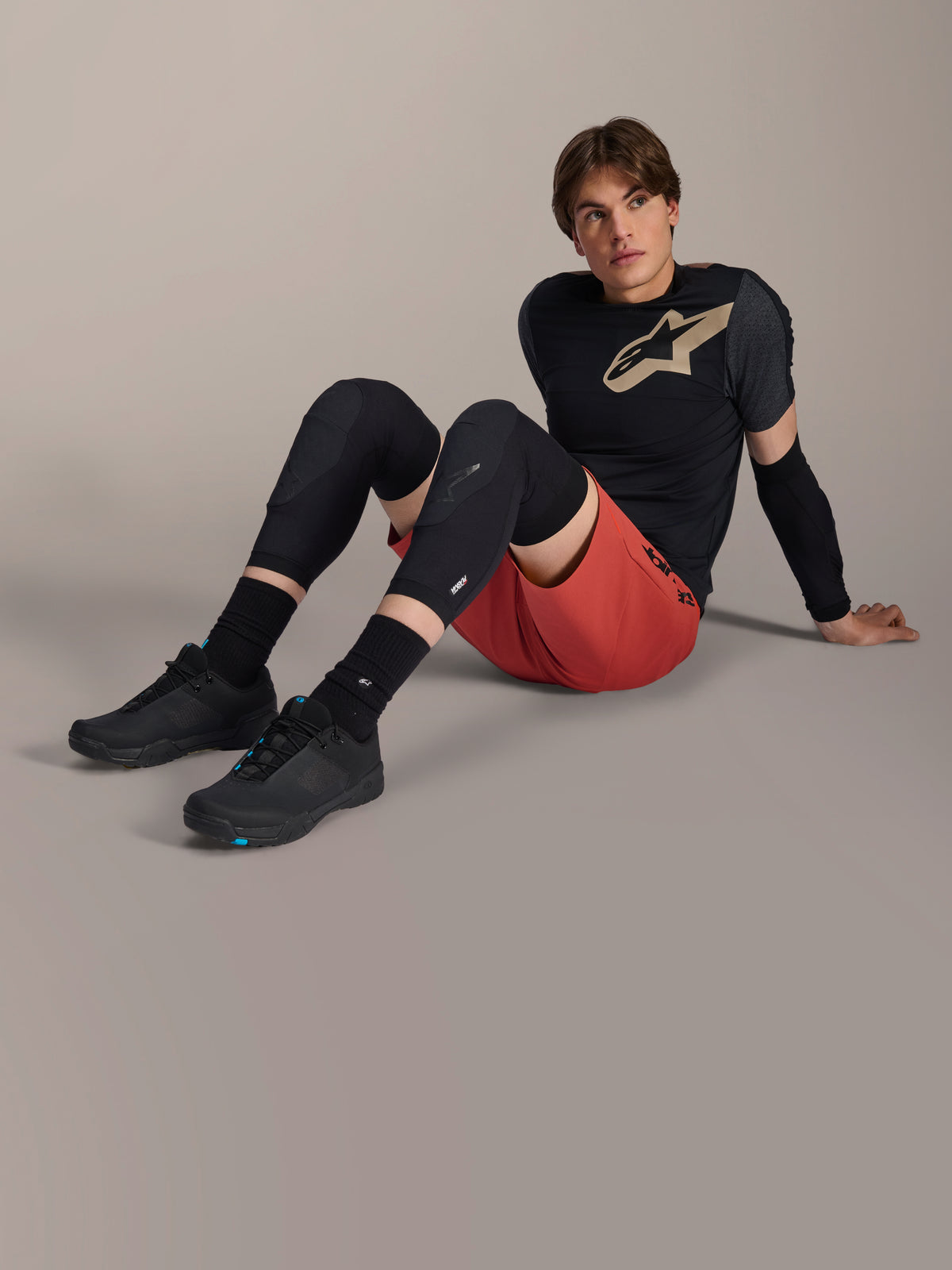 Alpinestars A-Motion Plasma Elbow Sleeve, worn by a male rider sitting on a studio floor, featuring black mesh construction, paired with red shorts and a black Alpinestars jersey with a tan logo, performance mountain bike protection for trail riding.
