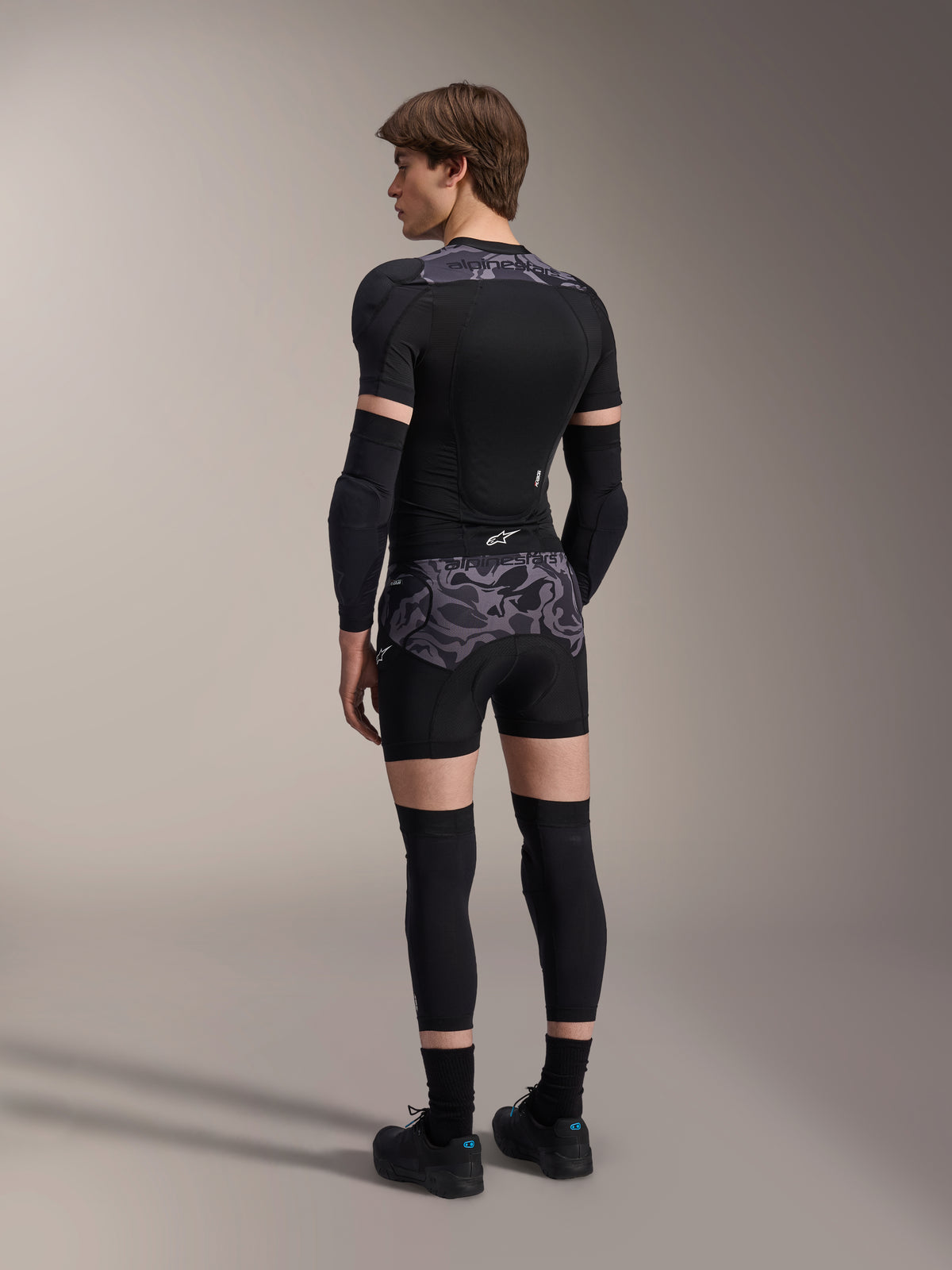 Model iført Alpinestars A-Motion Plasma Protection Boxers i Black Camo og matchende A-Motion Plasma Protection Jacket, set bagfra med gråt camouflage-mønster, albuebeskyttere, knæbeskyttere og cykelsko, komplet beskyttelsesudstyr til mountainbike.