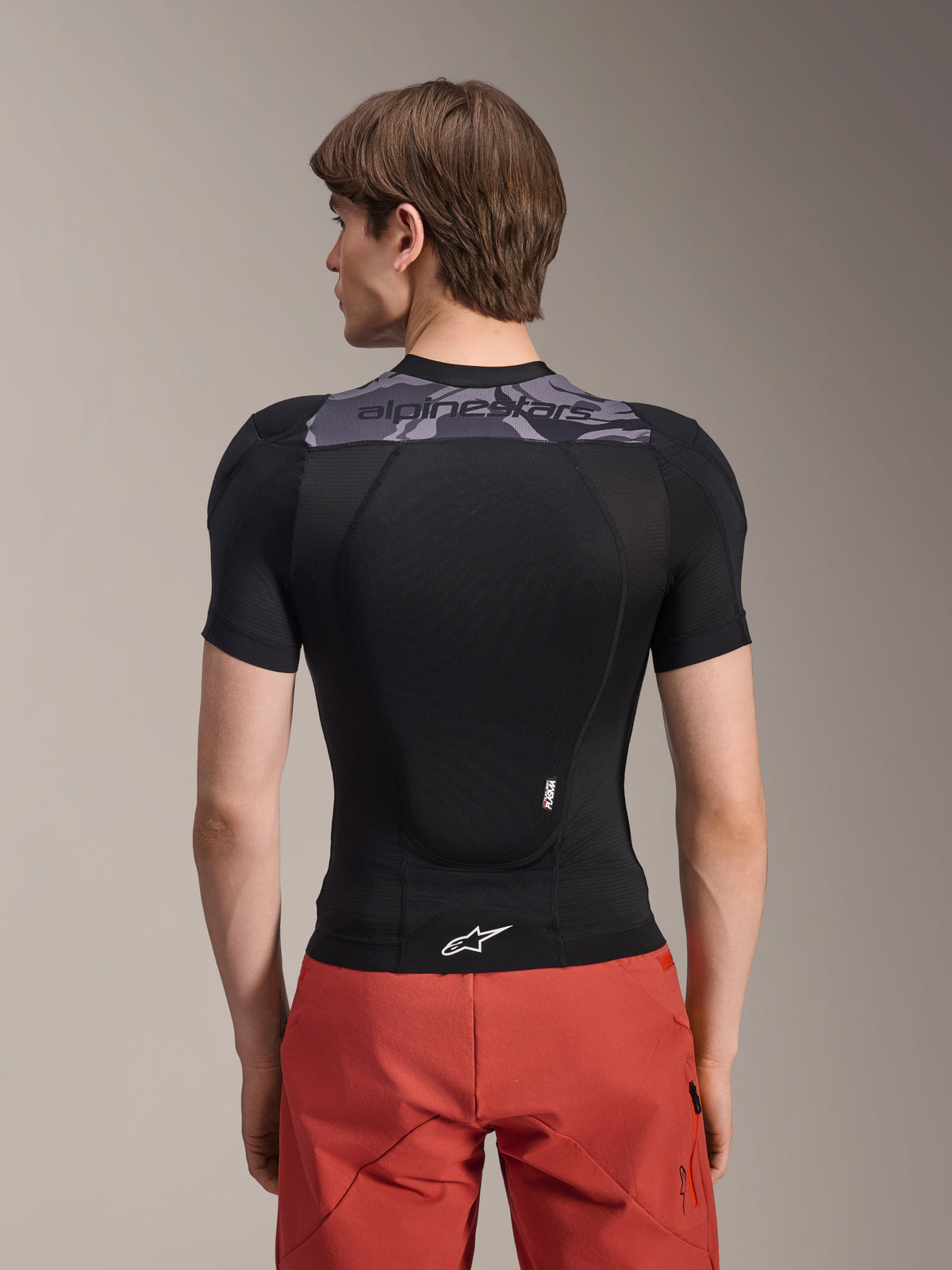 Alpinestars A-Motion Plasma beskyttelsesjakke - kortærmet, sort med grå camo-detaljer på den øverste del af ryggen, set bagfra med integreret rygskjold og hvidt Astars-logo ved kanten, kombineret med røde shorts, teknisk mountainbike-beskyttelsesudstyr
