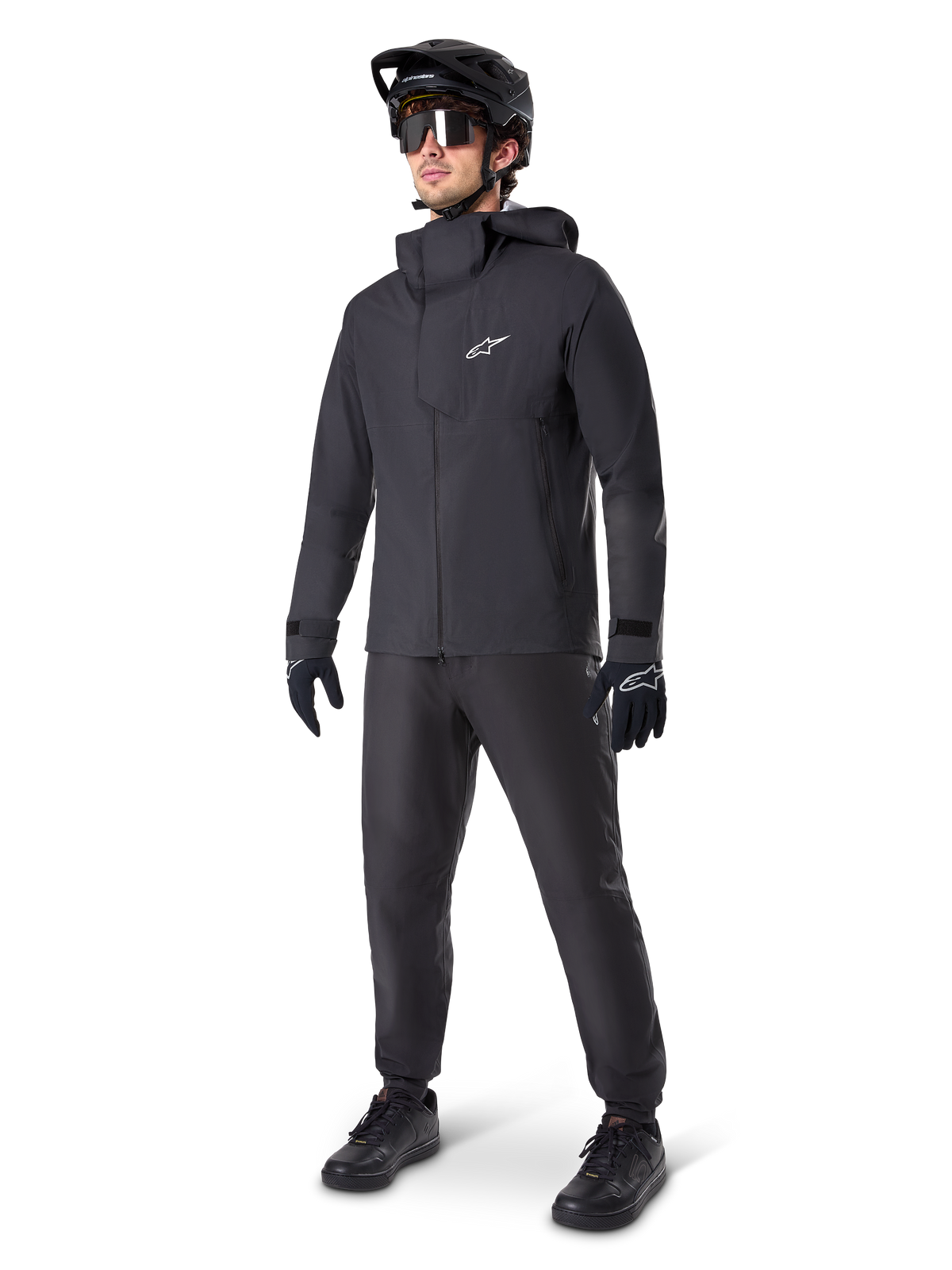 Mandlig mountainbiker iført Alpinestars A-Dura Waterproof Pants i sort, kombineret med en matchende sort vandtæt jakke med hætte, sort MTB-hjelm med visir, mørke cykelbriller og sorte handsker, komplet professionelt udstyrssæt til trail-kørsel i al slags vejr