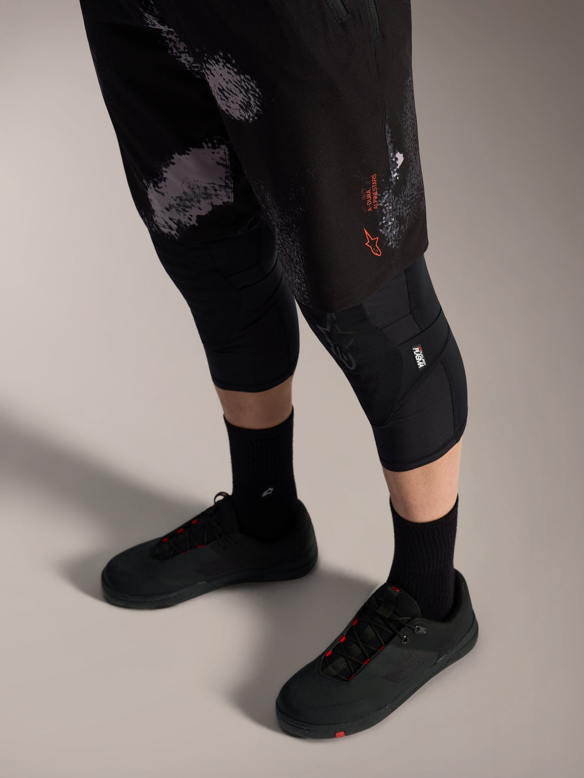 Alpinestars A-Dura Ride shorts i Black Camo med orange logo, vist med sorte knæbeskyttere og cykelsko til MTB