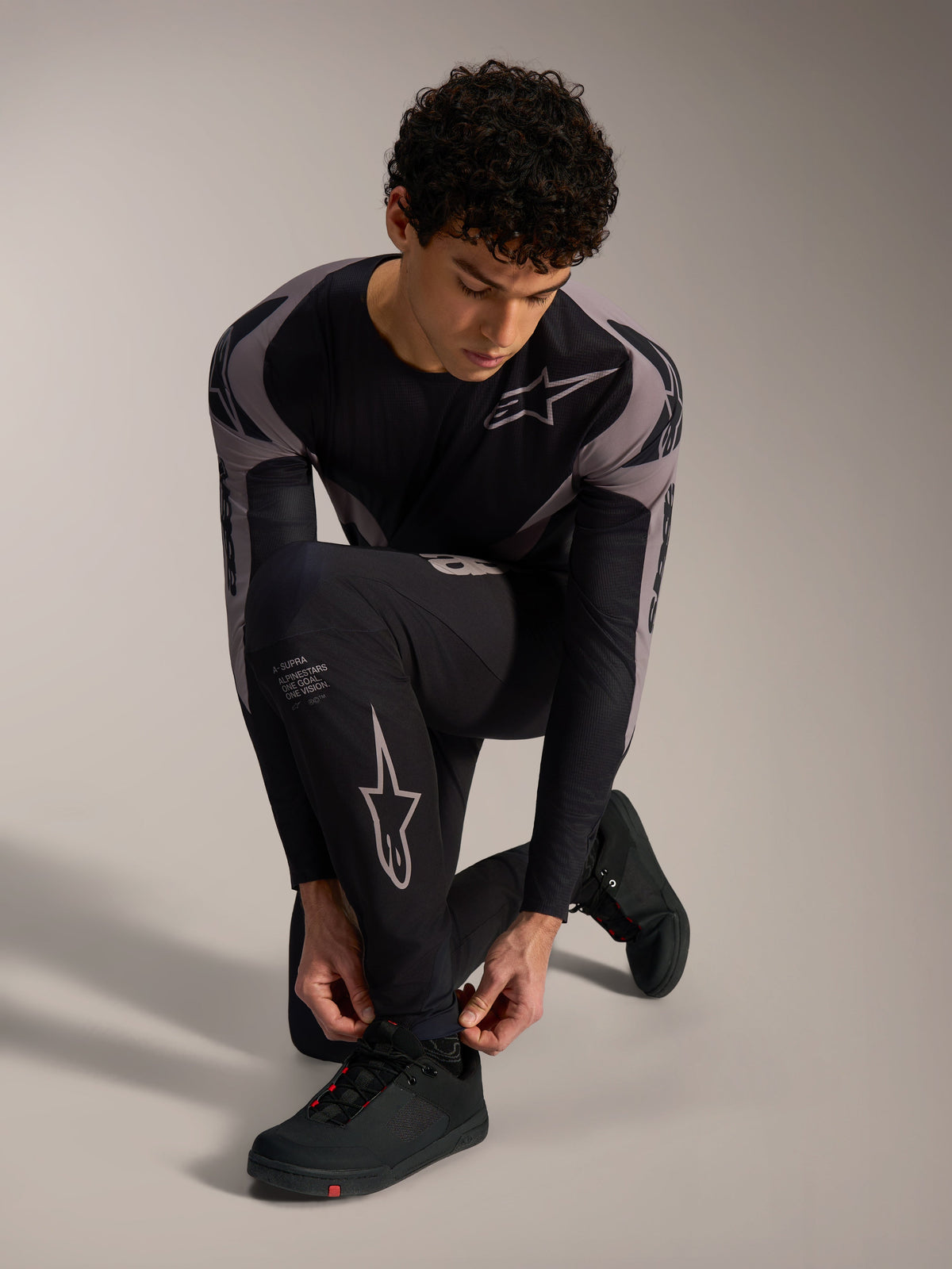 Sorte Alpinestars A-Supra Elite-bukser med grå racing-logoer, båret af en knælende model til MTB-kørsel.