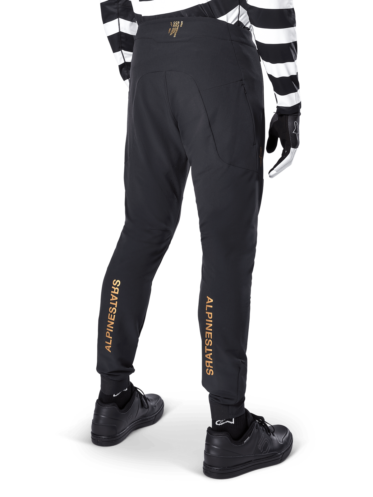 Alpinestars A-Aria Elite bukser, sorte MTB-bukser, set bagfra, med lodret guld Alpinestars-branding bag på benene, båret med sort- og hvidstribet trøje og sorte sko, performance-cykeltøj til mountainbike