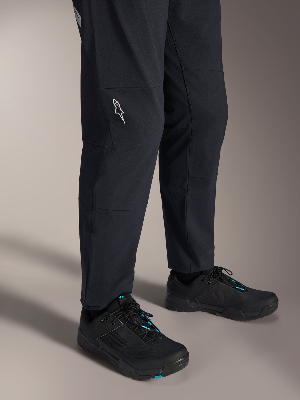 Alpinestars A-Dura Elite Pants, Black mountain bike pants, lower leg view featuring a white Alpinestars logo on the knee, worn with black and blue cycling shoes, durable construction for MTB riding