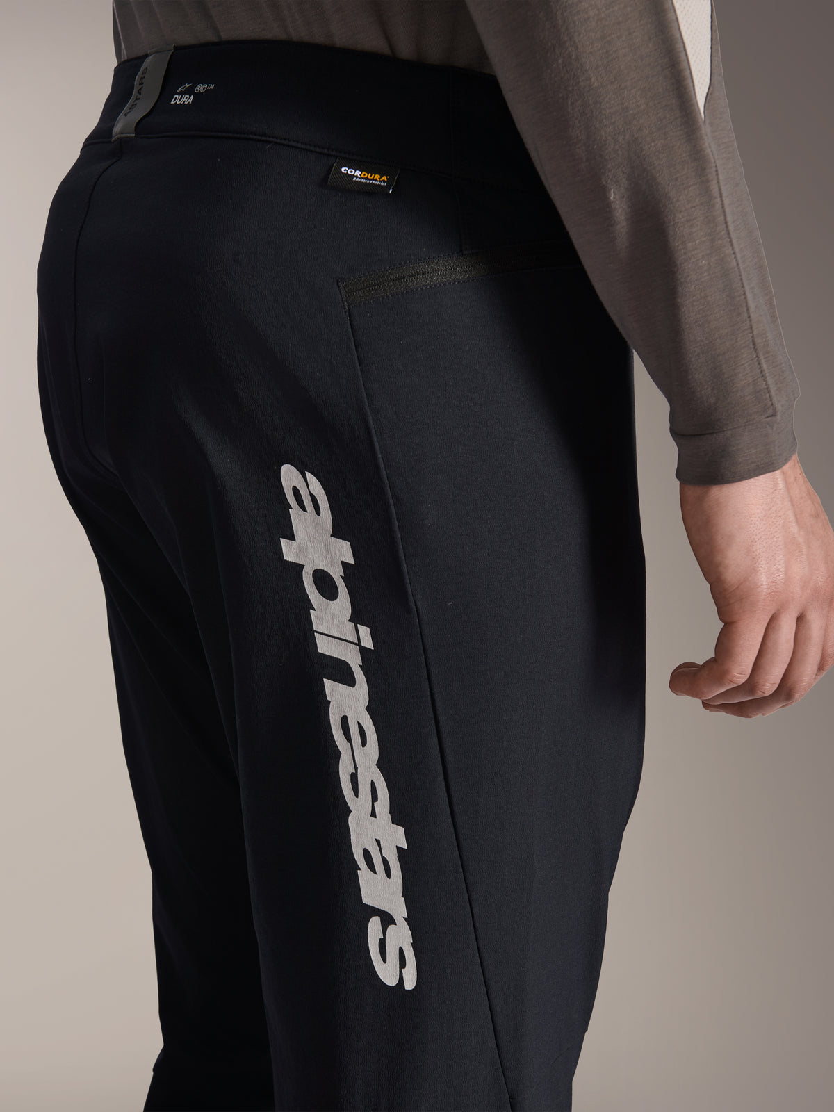 Alpinestars A-Dura Elite Pants, Black Mountain Bike Pants, side view featuring large vertical Alpinestars logo on the leg, Cordura fabric tag, and zippered lateral pocket, technical cycling apparel for off-road riding.