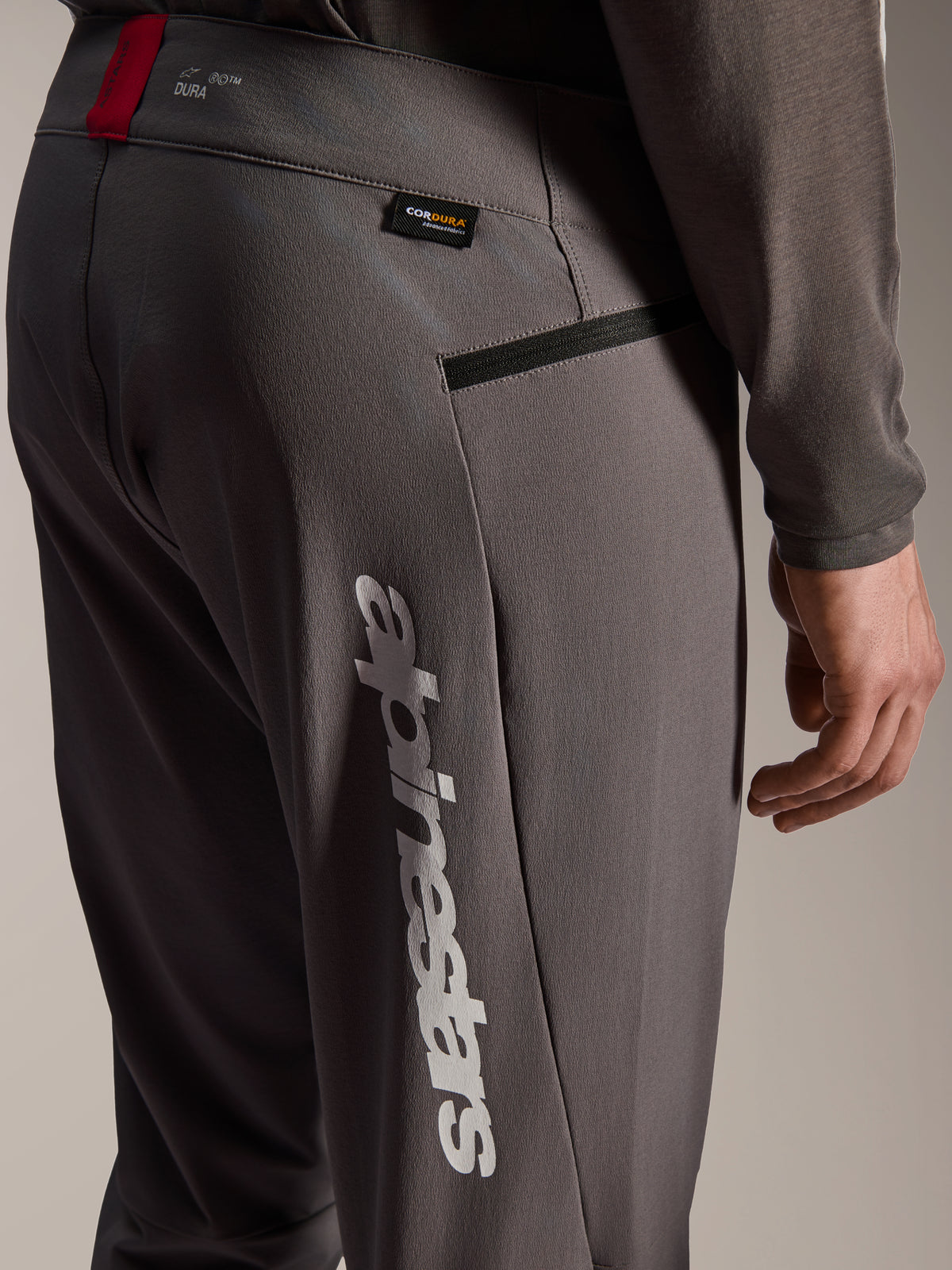 Alpinestars A-Dura Elite bukser, mørkegrå MTB-bukser, nærbillede af hofte og lår, med stort hvidt lodret Alpinestars-logo, Cordura-stofmærke, lynlåslomme og rød detalje på linningen