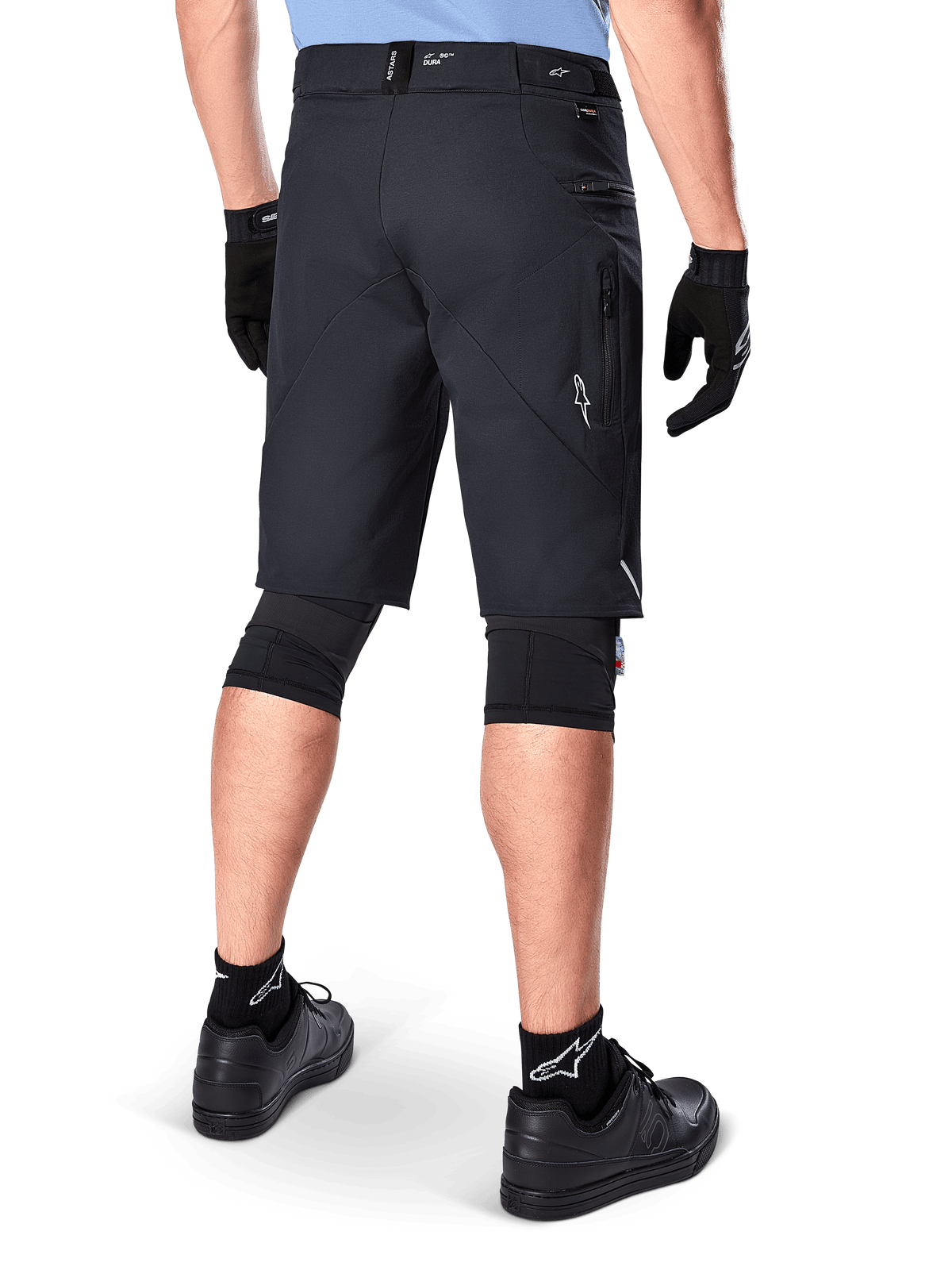 Alpinestars A-Dura Elite Shorts, sort, set bagfra af en rytter iført mountainbike-udstyr inklusiv sorte knæbeskyttere, handsker og strømper med hvide logoer, med lynlåslommer i siden og reflekterende detaljer på slidstærkt Cordura-stof.