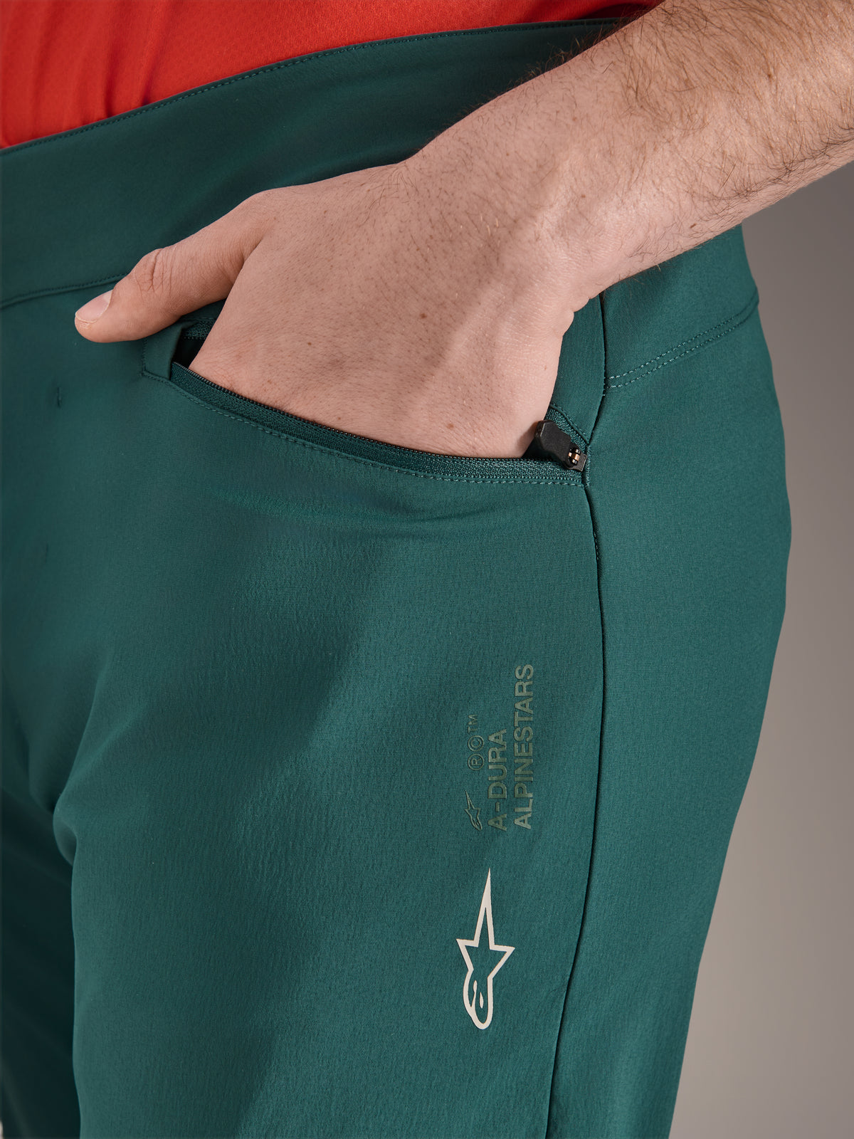 Alpinestars A-Dura bukser i Dark Pine grøn, tætbillede af lynlåslomme med en hånd, hvidt Alpinestars-logo og tekst på låret, slidstærke MTB-cykelbukser