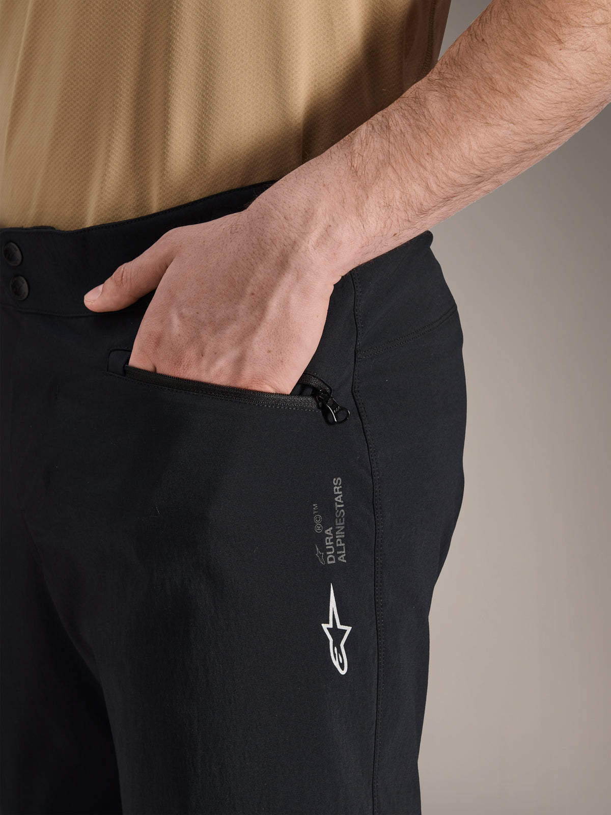 Alpinestars A-Dura shorts, sorte mountainbike-shorts, nærbillede af sidelomme med lynlås og en hånd indeni, med hvidt Alpinestars-logo og A-Dura-branding, slidstærkt tekstil til off-road cykling