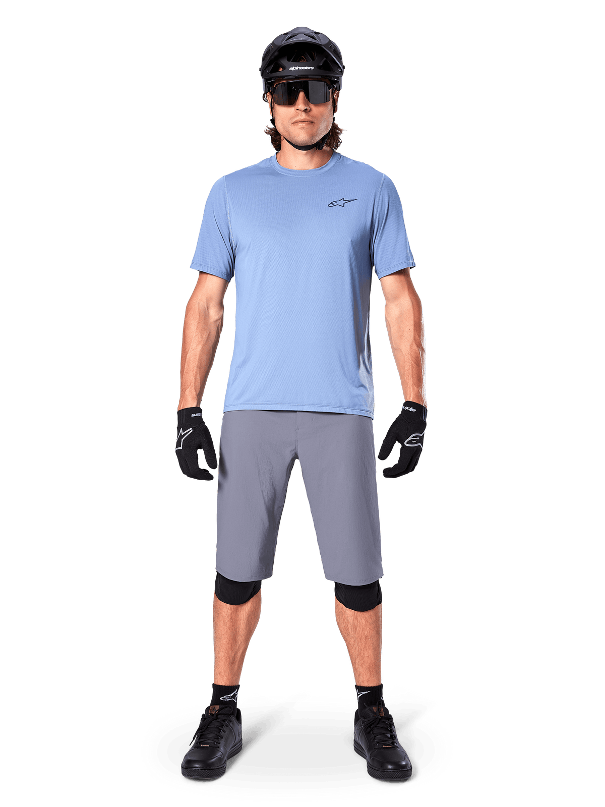 Mandlig rytter iført Alpinestars A-Dura Shorts i mørkegrå, kombineret med en lyseblå kortærmet t-shirt, sort hjelm, solbriller og sorte knæbeskyttere, komplet mountainbike-outfit til terrænkørsel