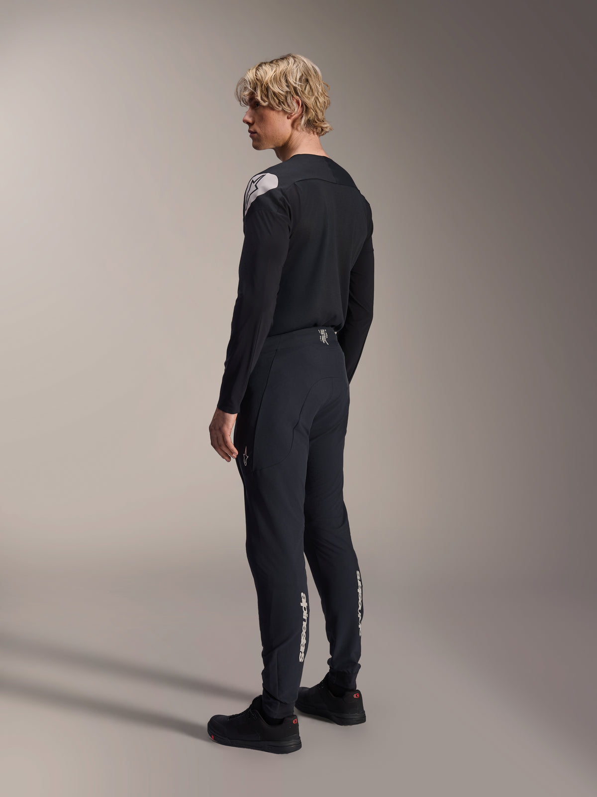 Alpinestars A-Aria Elite-bukser, mountainbike-bukser, Black Titanium, trekvart-visning bagfra på model, med hvidt Alpinestars-branding på underbenene, performance-pasform med elastiske manchetter, kombineret med en matchende sort langærmet jersey.