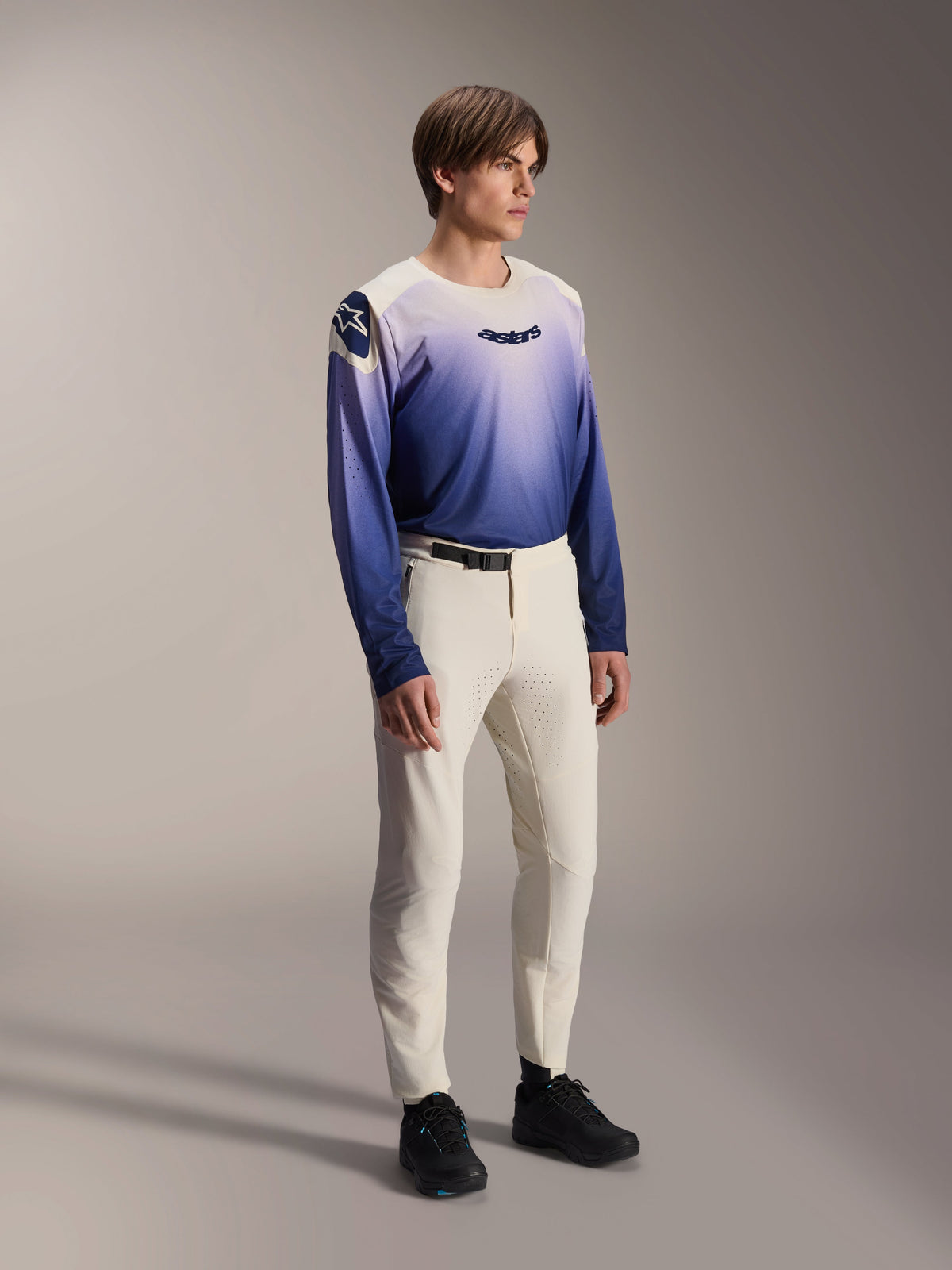 Male model wearing the Alpinestars A-Aria Polartec Maker Elite Jersey in Baja Blue, featuring a white-to-blue gradient design with a black 'astars' logo on the chest, paired with off-white mountain bike pants and black shoes, performance-fit MTB apparel for trail riding.