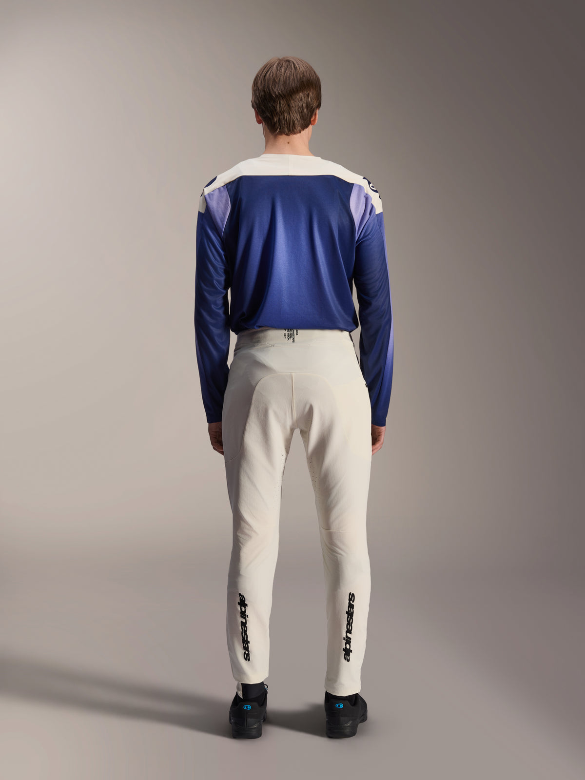 Alpinestars A-Aria Elite-bukser i Ecru, vist bagfra på en model kombineret med en blå A-Aria Polartec Maker Elite langærmet jersey, råhvide tekniske MTB-bukser med sorte logoer på underbenene, performance-cykeludstyr til mountainbike