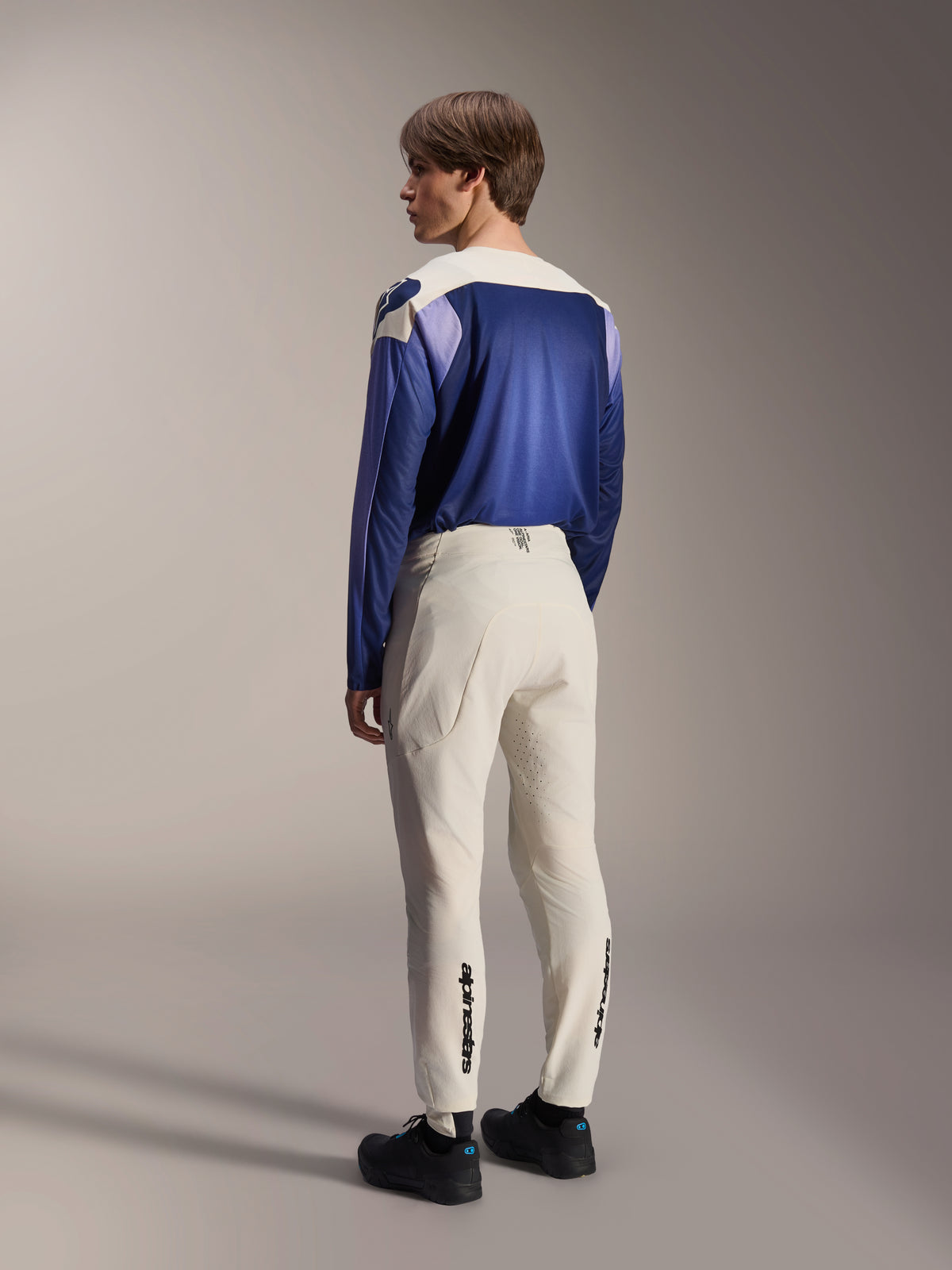 Alpinestars A-Aria Elite bukser, MTB-bukser, Ecru off-white, set bagfra på en model, med sort tekst-logo på underbenene og laserperforeringer til ventilation, kombineret med en blå og lilla langærmet trøje og sorte sko, professionelt mountainbike-udstyr