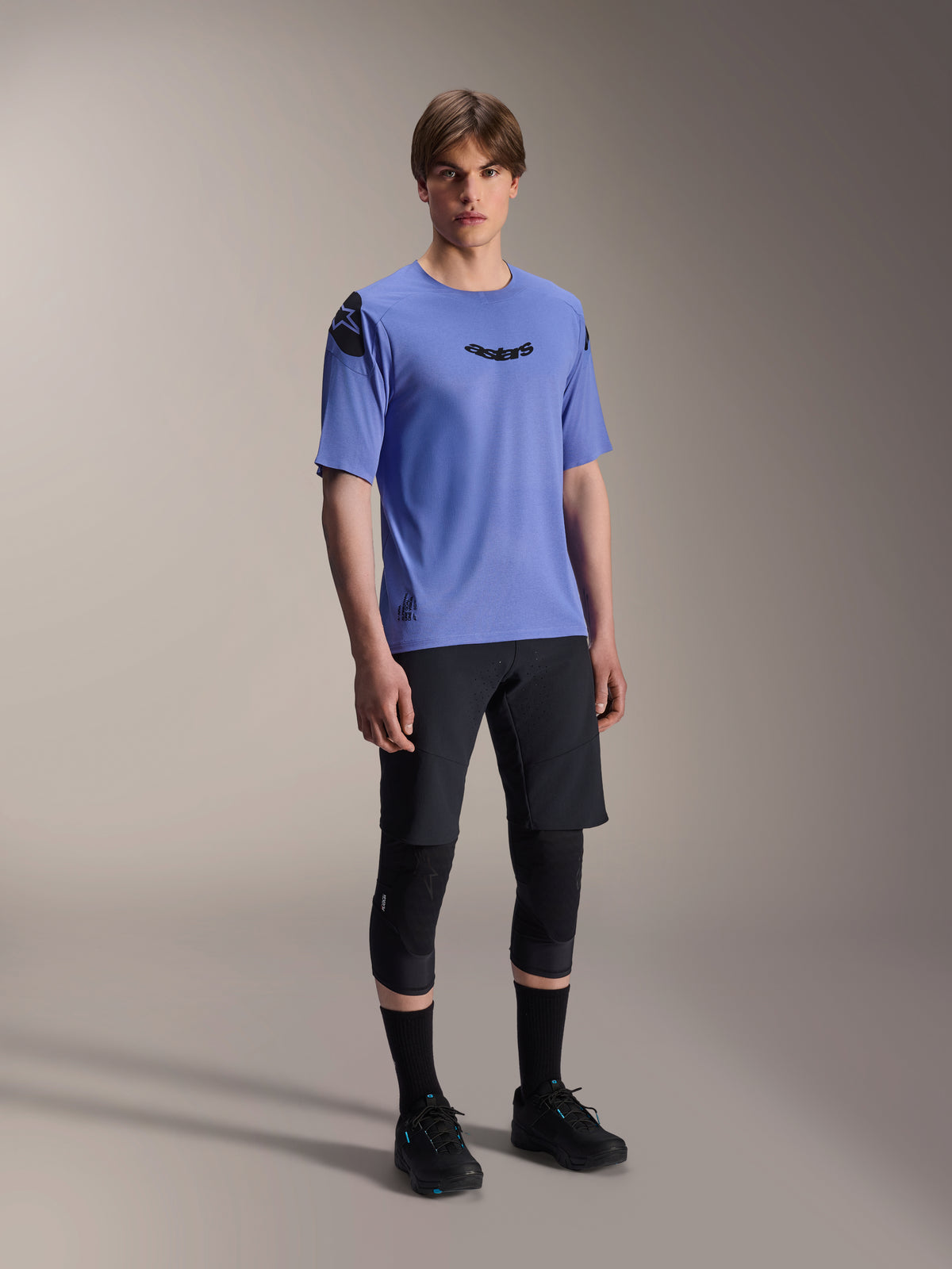 Mandlig model iført Alpinestars A-Aria Lite-shorts i sort, kombineret med en blå A-Aria Polartec Elite kortærmet trøje, sorte knæbeskyttere og sorte cykelsko, performance mountainbike-beklædning til trail-kørsel.