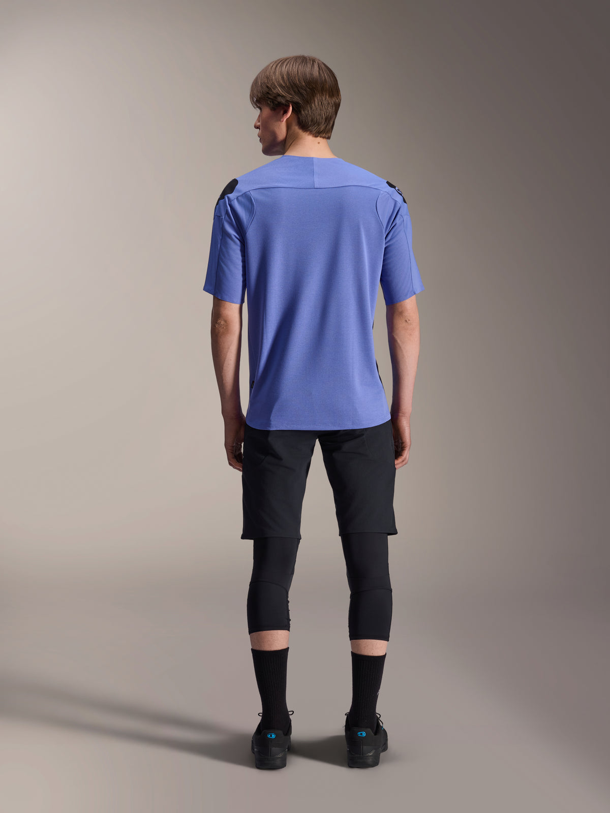 Mountainbikerytter set bagfra iført Alpinestars A-Aria Lite Shorts i sort, kombineret med en blå kortærmet jersey, sorte knæbeskyttere og sorte strømper, performance-cykeludstyr til off-road trails