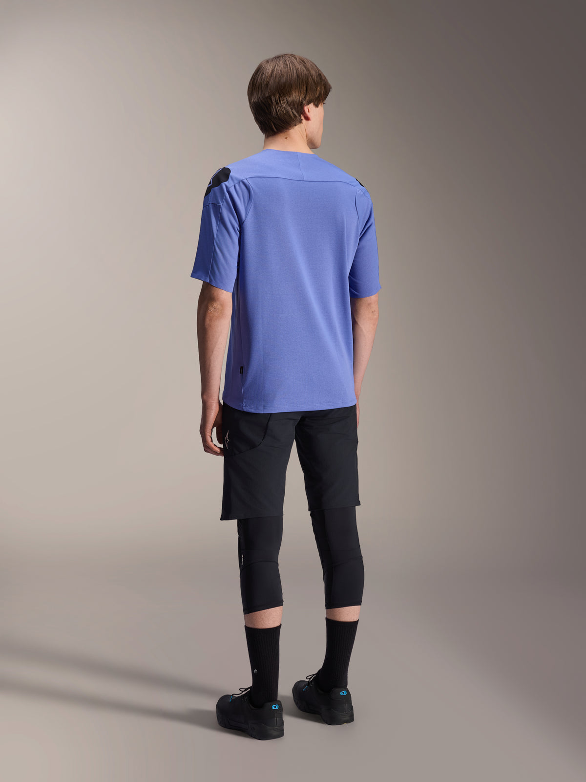 Alpinestars A-Aria Lite shorts, sorte mountainbike-shorts, kombineret med en blå A-Aria Polartec Elite kortærmet trøje og sorte knæbeskyttere, set bagfra af en rytter i performance-cykeludstyr til off-road stier.