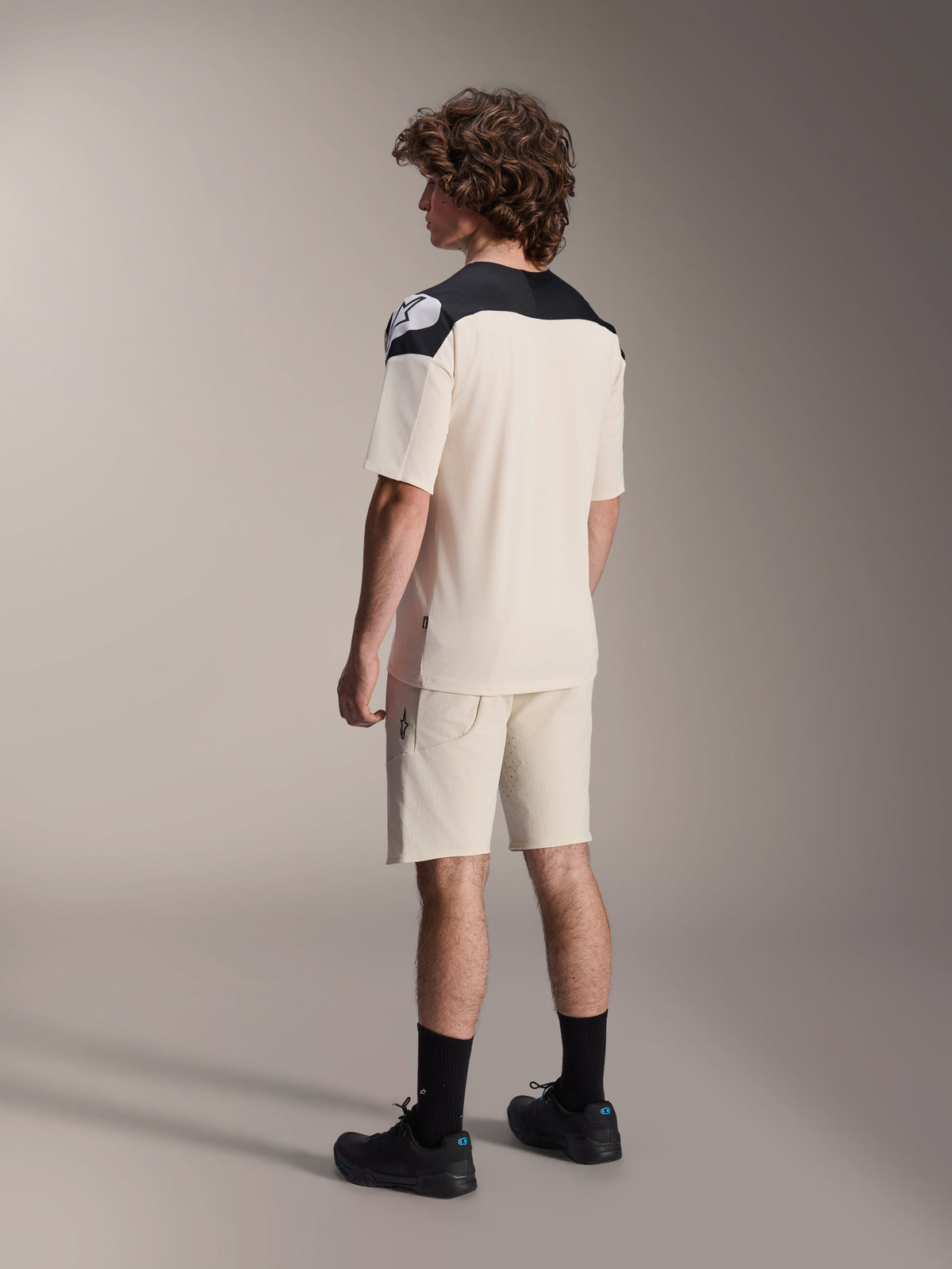 Model wearing the Alpinestars A-Aria Polartec Elite Short Sleeve Jersey in Ecru, viewed from behind, featuring black shoulder panels with a white logo, styled with matching ecru mountain bike shorts and black shoes