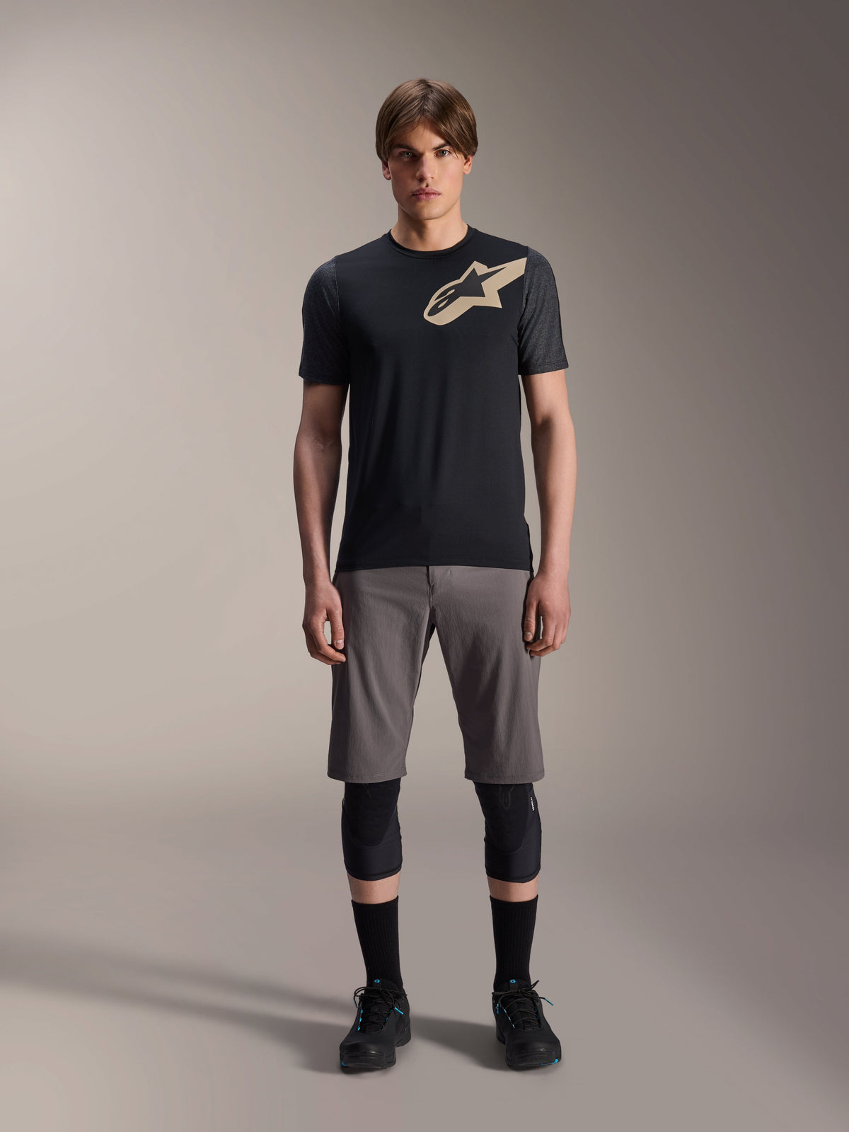 Alpinestars A-Dura Lipan kortærmet jersey, sort med gråmêlerede ærmer og tan-farvet Astars-logo på brystet, båret af en mandlig model med grå mountainbike-shorts og sorte knæbeskyttere, performance MTB-køreudstyr