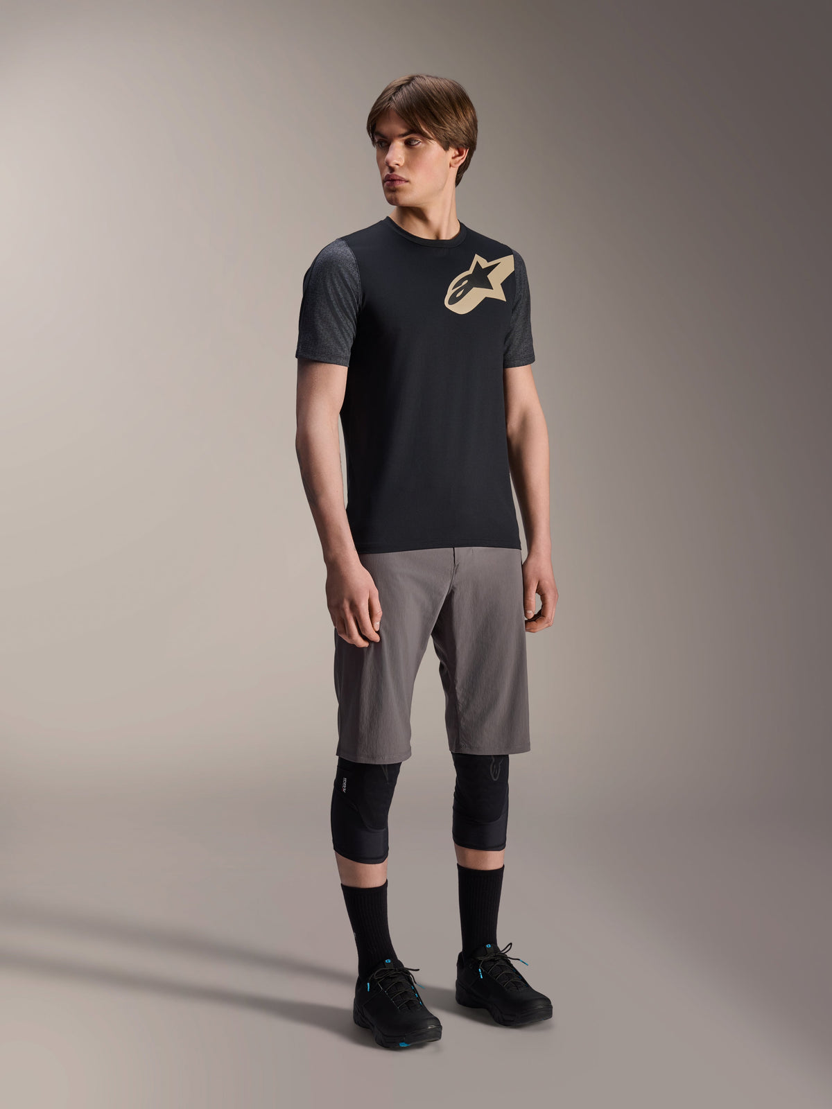 Alpinestars A-Dura Lipan kortærmet jersey i sort, mørkegrå ærmer og et tan Astars-logo på brystet, kombineret med grå mountainbike-shorts, sorte knæbeskyttere og sorte cykelsko, koordineret mountainbike-udstyr