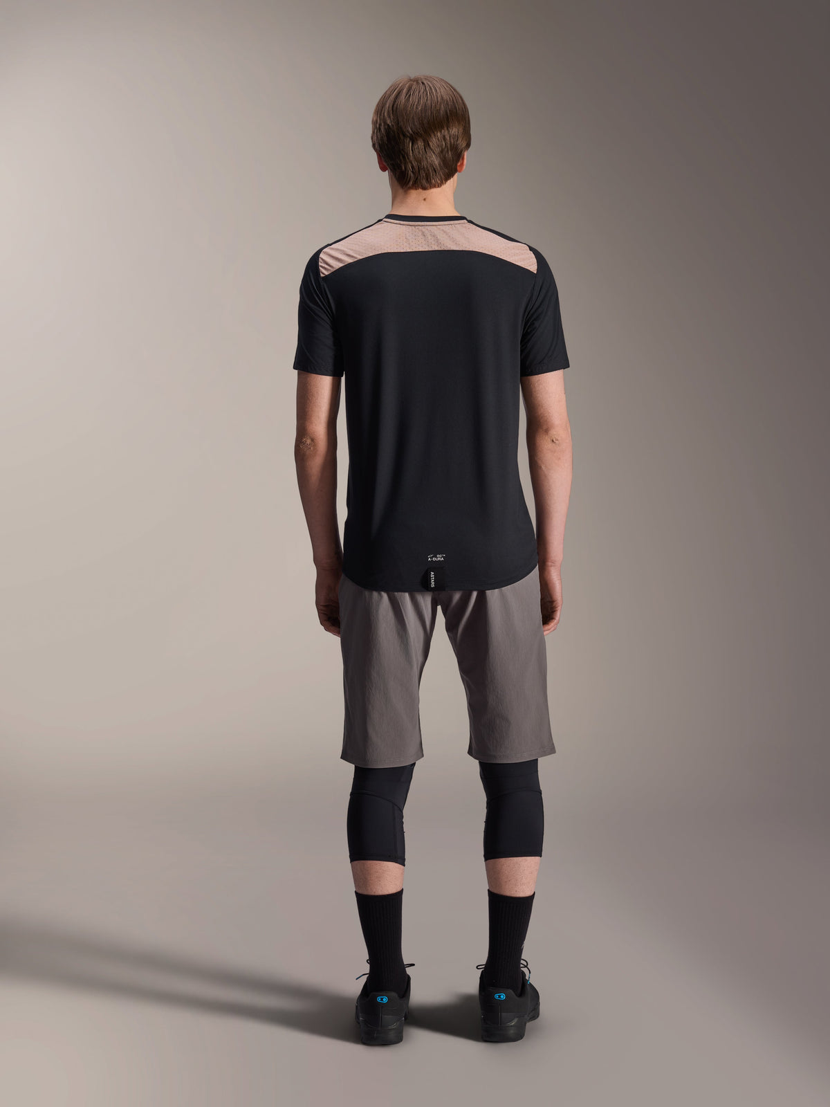 Alpinestars A-Dura Lipan Jersey - Kortærmet, sort med lysebrunt øvre rygpanel, set bagfra med forlænget rygstykke, båret med grå MTB-shorts og sorte knæbeskyttere, performance mountainbike-beklædning