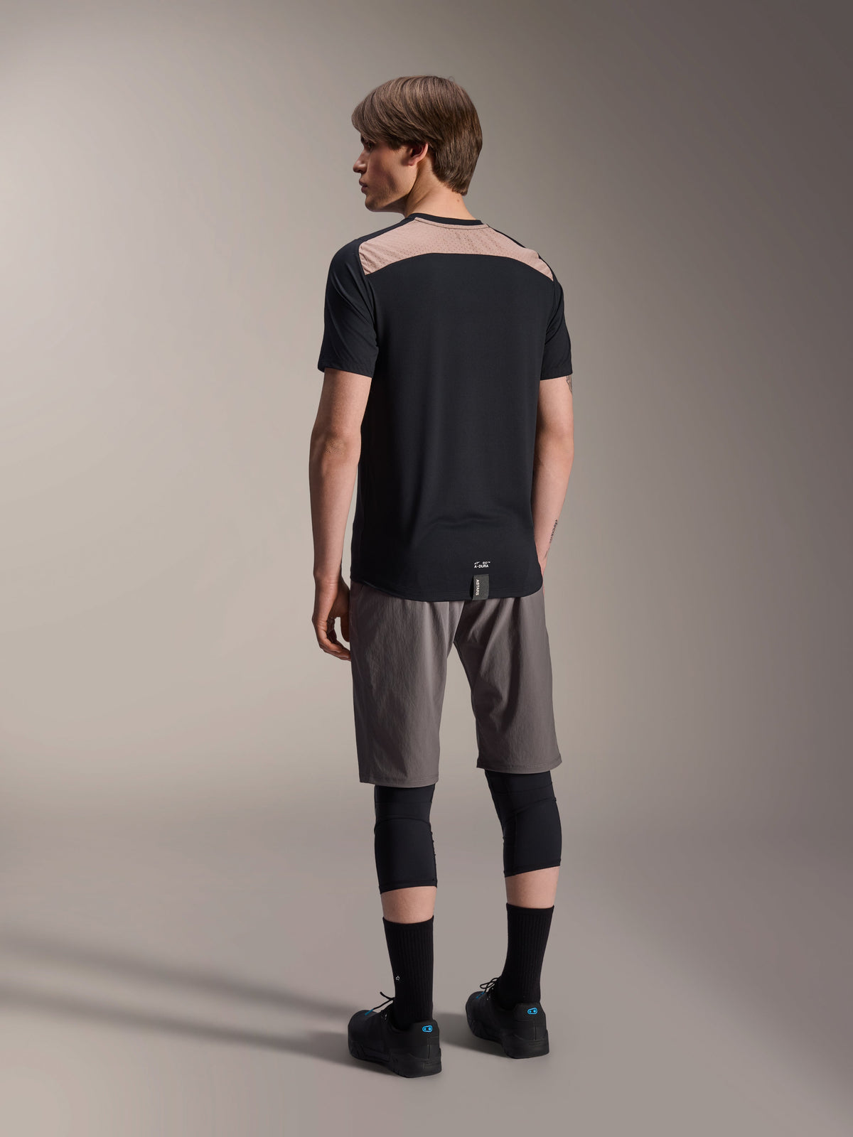 Alpinestars A-Dura Lipan kortærmet jersey i sort, set bagfra på model, der viser det lysebrune perforerede mesh-panel over den øverste del af ryggen, kombineret med grå mountainbike-shorts og sorte knæbeskyttere for et komplet trail-outfit.