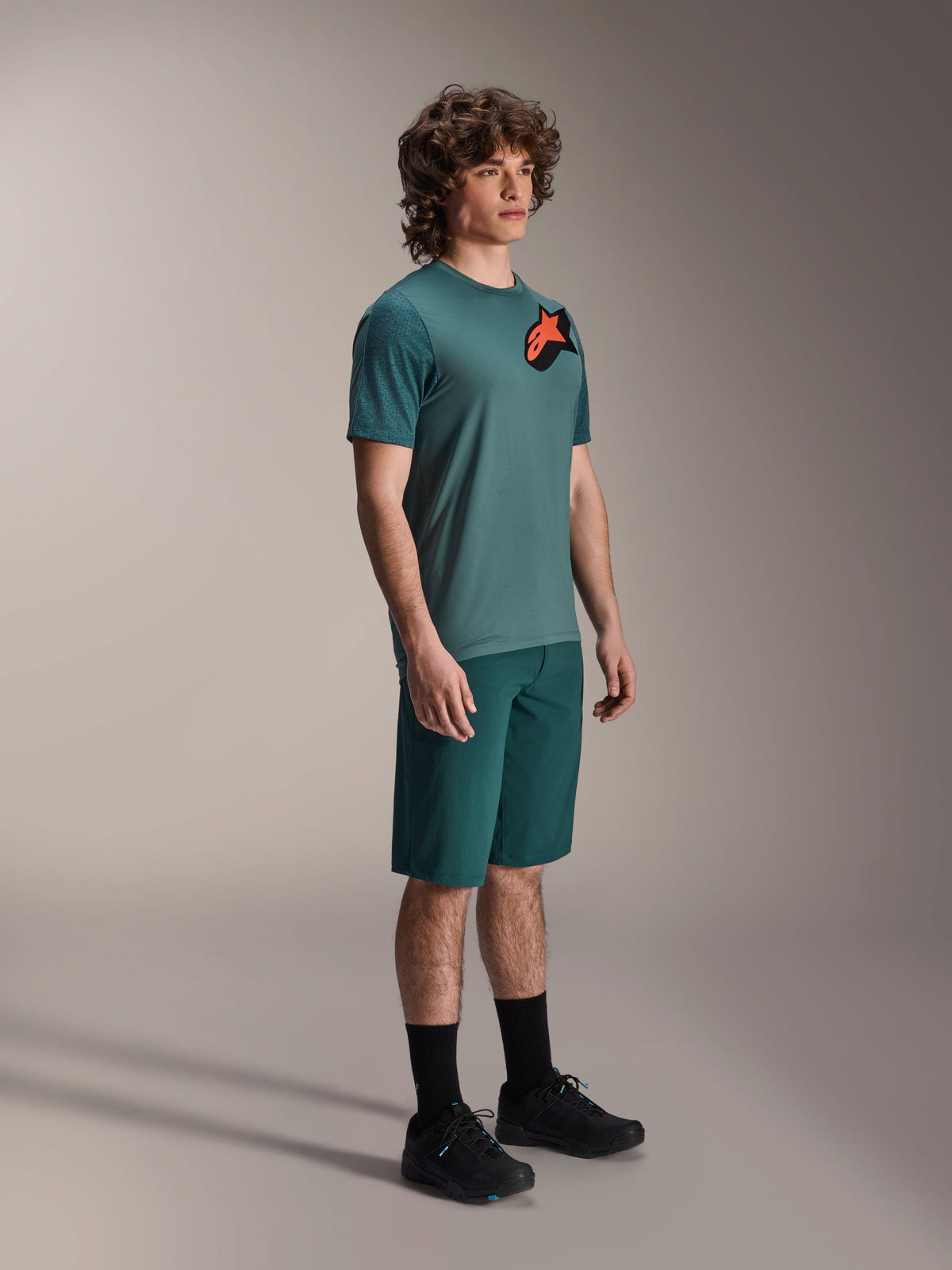Alpinestars A-Dura Lipan jersey, kortærmet MTB-trøje, Silver Pine grøn, med orange og sort Astars-logo på brystet, kombineret med matchende grønne shorts og sorte cykelsko, afslappet trail-stil