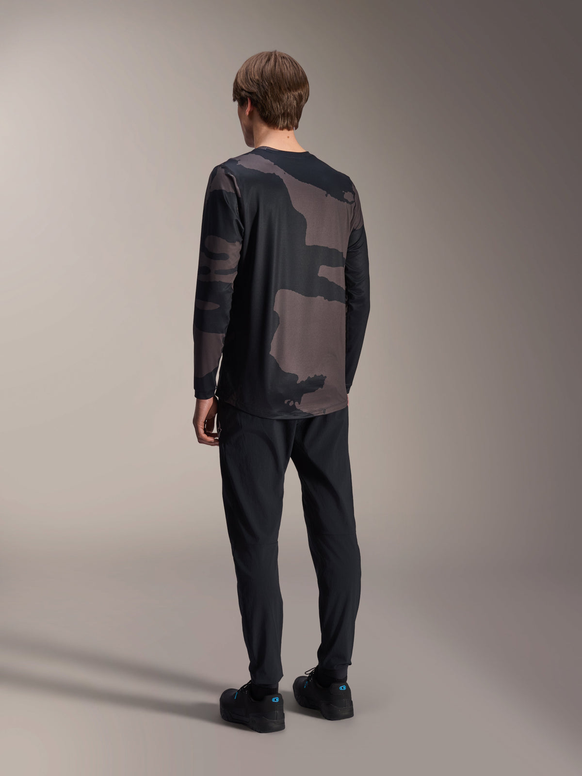 Alpinestars A-Dura Hollow Camo Long Sleeve Jersey, Black Camo, rear view of model, technical mountain bike jersey with dropped tail hem, paired with black pants and cycling shoes, casual off-road performance gear