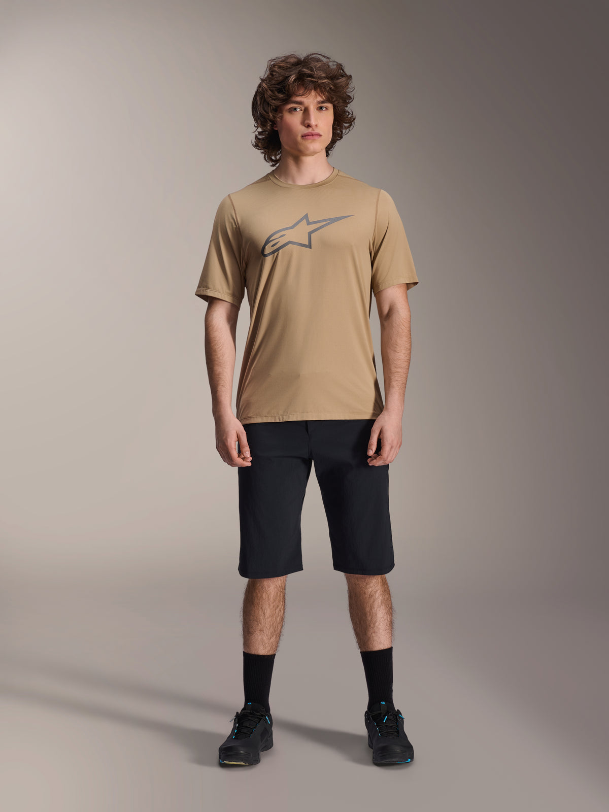Model iført Alpinestars A-Dura SS Jersey i beige og A-Dura Shorts i sort, med et gråt stjernelogo på brystet, kombineret med sorte strømper og mountainbike-sko, komplet MTB-beklædning i studieomgivelser.