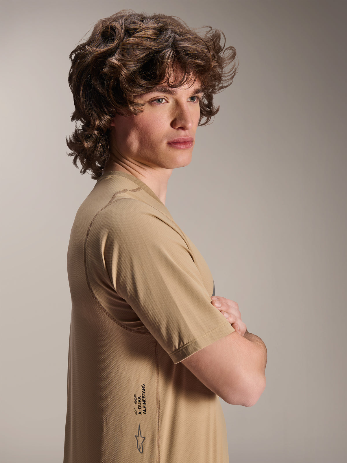 Alpinestars A-Dura Astar Jersey, Short Sleeve MTB Jersey, Light Brown, side view of a model with wavy hair wearing the textured jacquard jersey, featuring the A-Dura logo on the lower side, casual mountain bike performance style