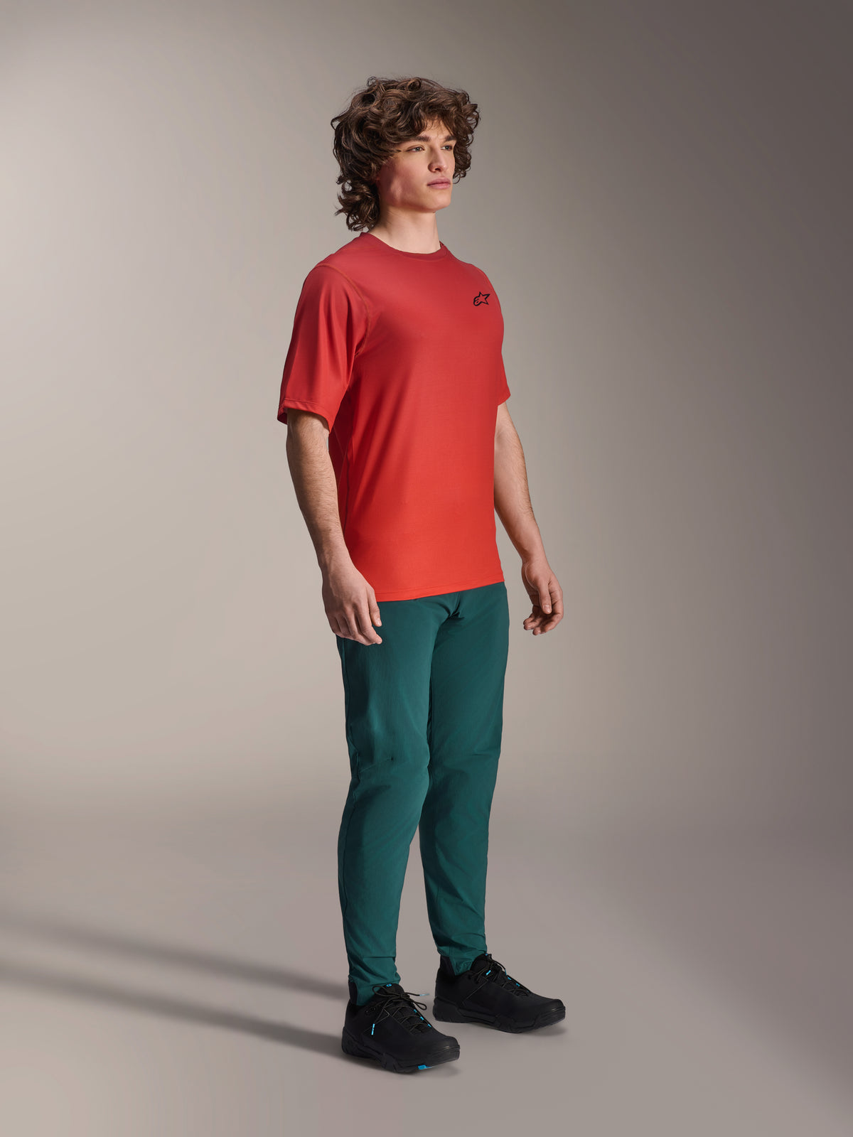 Model iført Alpinestars A-Dura bukser i Dark Pine, kombineret med en rød A-Dura Rise kortærmet jersey og sorte MTB-sko, atletisk mountainbike-beklædning i et studiemiljø