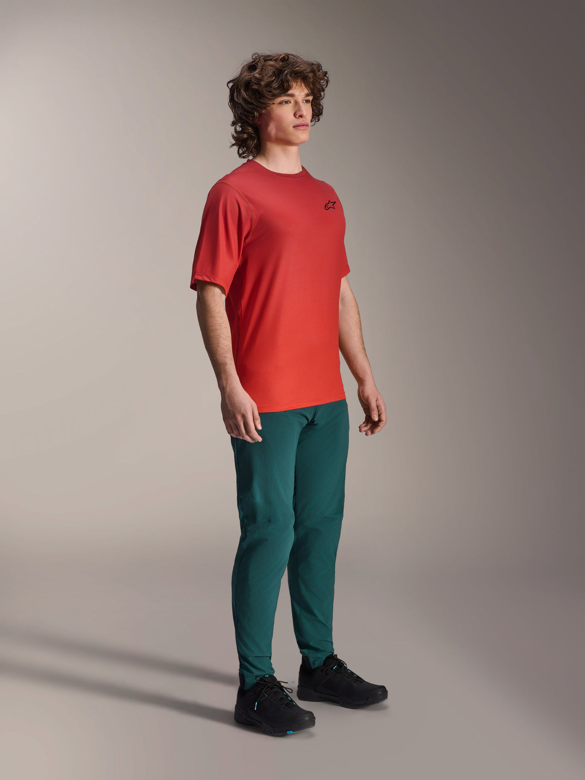 Alpinestars A-Dura Rise jersey, kortærmet, Spicy Orange, med et lille sort logo på brystet, båret med mørke blågrønne bukser og sorte sko, letvægts fugttransporterende mountainbike-beklædning