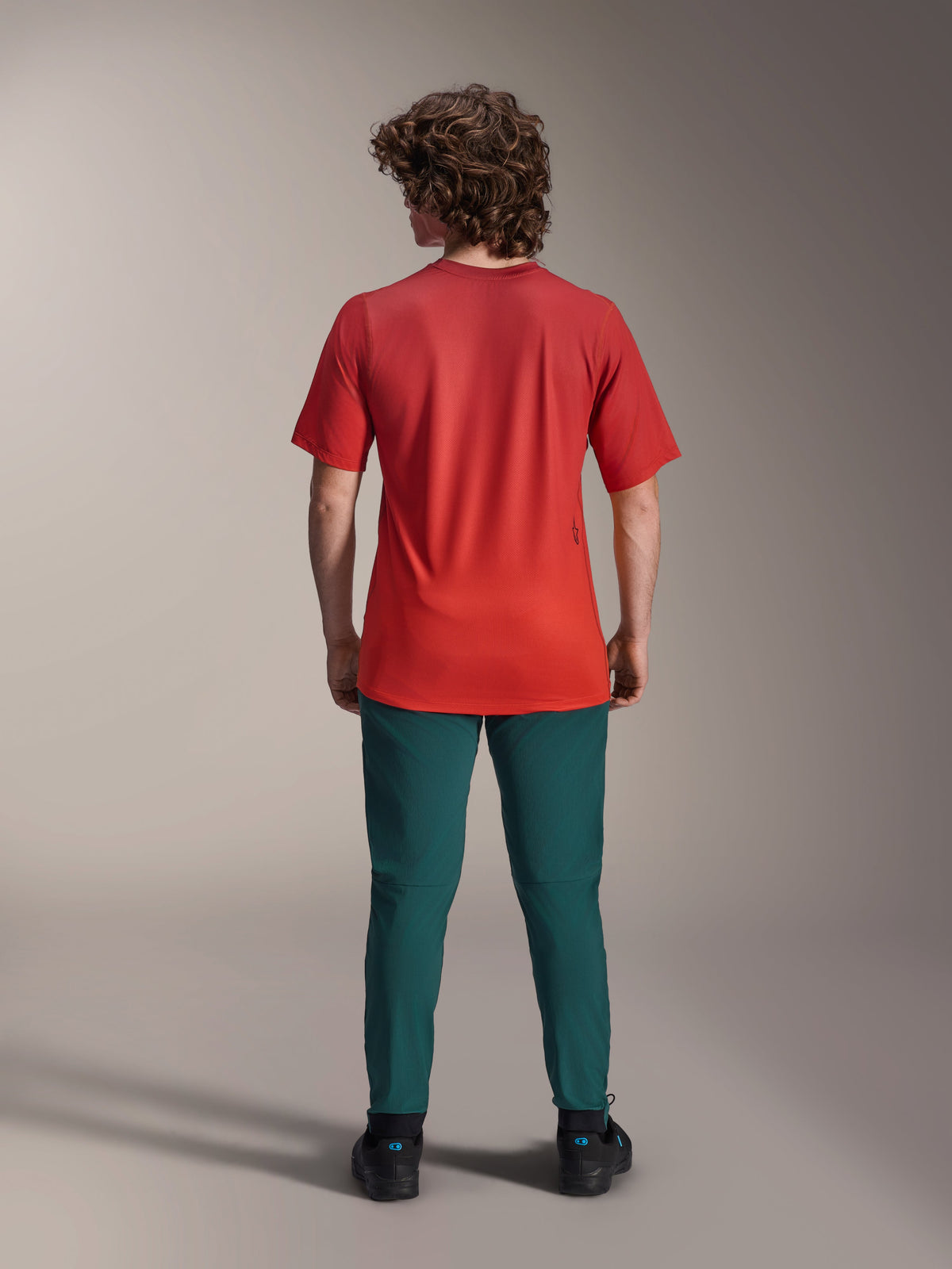 Alpinestars A-Dura Rise kortærmet jersey, Spicy Orange, set bagfra på en model iført mørke petroliumsblå bukser og sorte sko, med forlænget bagkant og et lille skærmtrykt logo på ryggen, letvægts performance MTB-udstyr