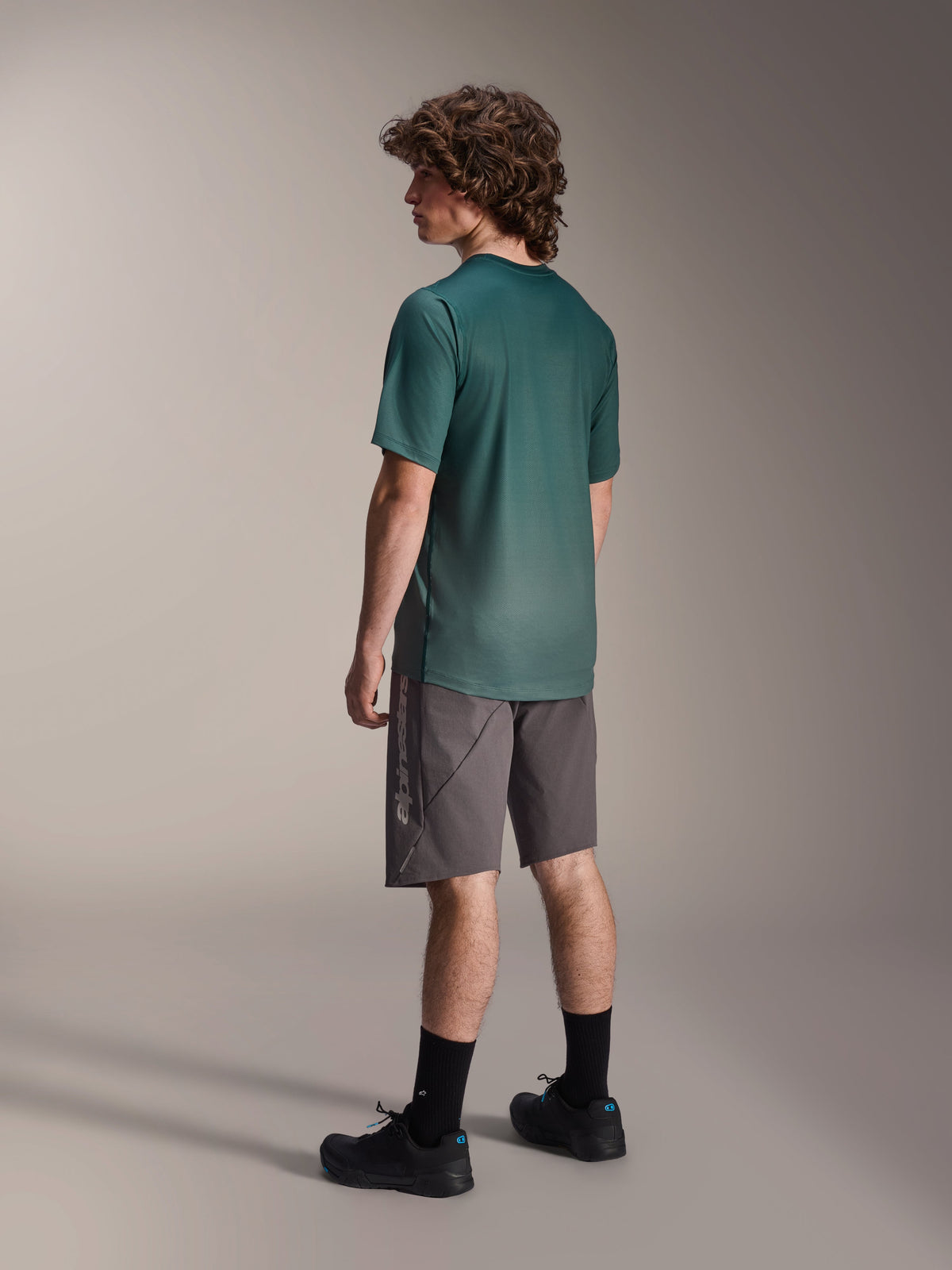 Alpinestars A-Dura Rise Jersey, kortærmet MTB-trøje, Silver Pine Green, båret med grå shorts og sorte sko, set bagfra med længere drop tail og almindelig pasform, letvægts fugttransporterende materiale til trailkørsel