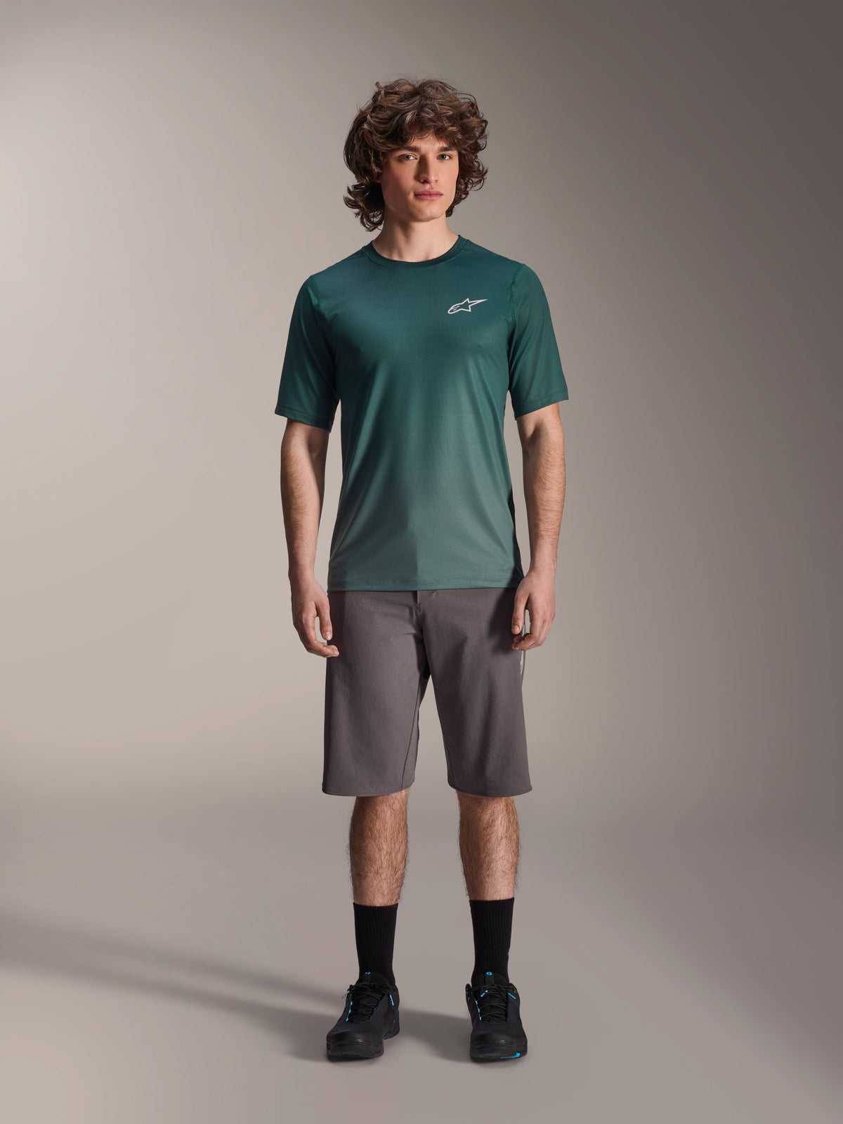 Mandlig model i Alpinestars A-Dura Rise Jersey - Kortærmet i Silver Pine, grøn cykeltrøje med lille hvidt logo på brystet, kombineret med grå mountainbike-shorts og sort fodtøj, afslappet MTB-trailudstyr