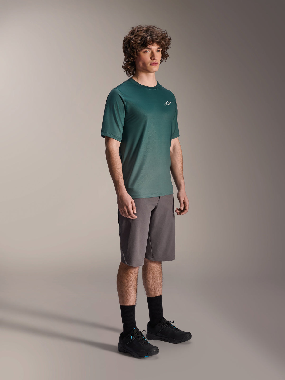 Alpinestars A-Dura Rise Jersey - kortærmet i Silver Pine, grøn mountainbike-jersey båret af en model med grå shorts og sorte sneakers, afslappet MTB-udstyr til trailkørsel, lille hvidt logo på brystet