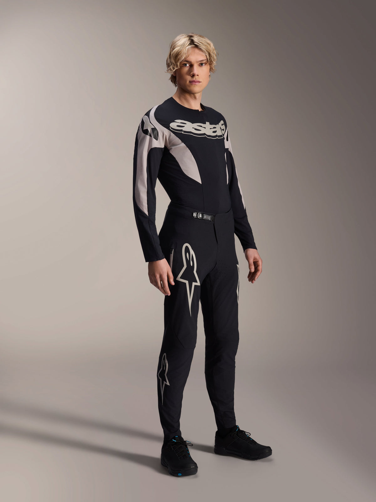 Mandlig model iført Alpinestars A-Supra Melt-bukser og matchende langærmet jersey, sort med lysegrå buede mønstre, markante hvide Astars-logoer på benene og ordmærke på brystet, professionelt mountainbike racing-sæt