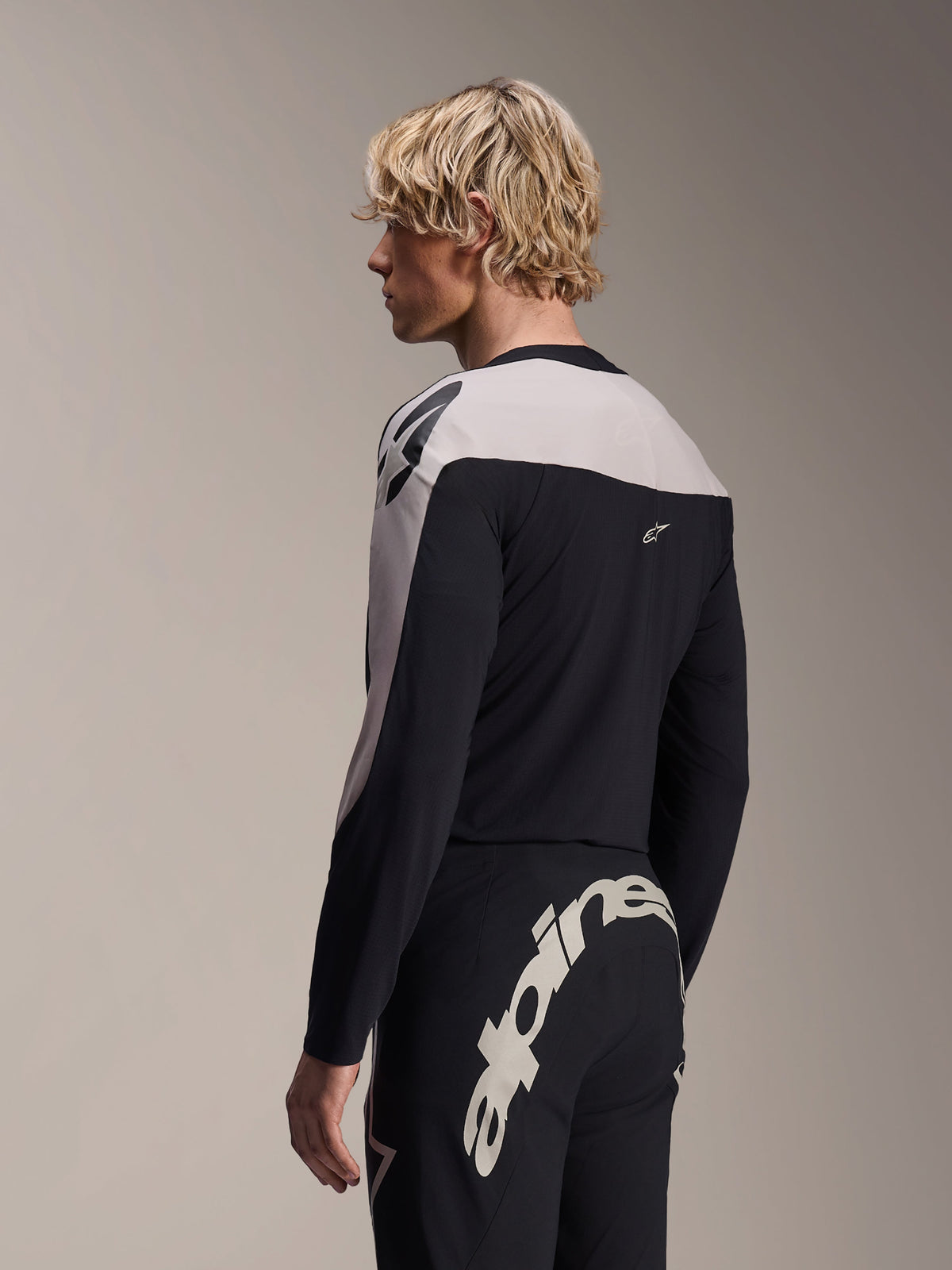 Mandlig model iført Alpinestars A-Supra Melt langærmet jersey og A-Supra Melt bukser, sort og lysegråt MTB-racersæt, med et stort hvidt Alpinestars-ordmærke på tværs af buksernes bagstykke.