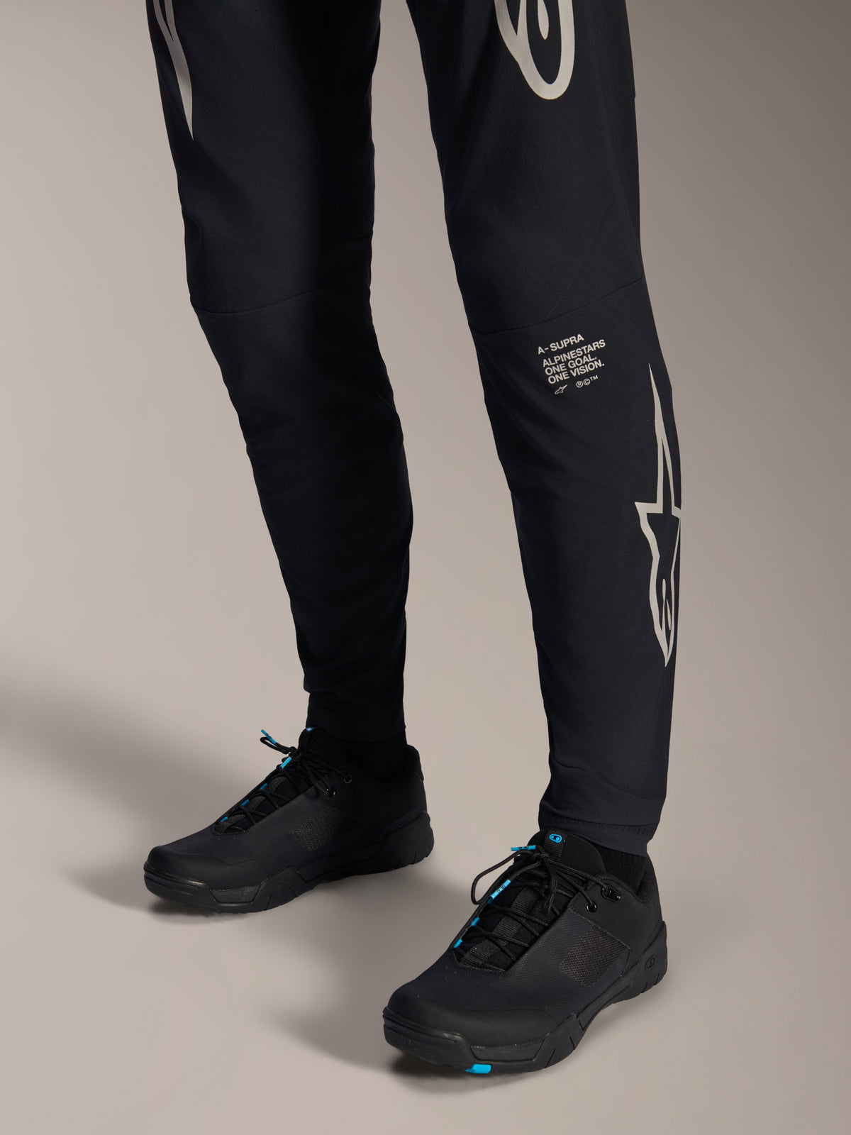 Alpinestars A-Supra Melt bukser, sorte mountainbike-bukser, med hvidt Alpinestars ordmærke og logo på benet, båret med sorte cykelsko med blå detaljer, performance off-road-udstyr