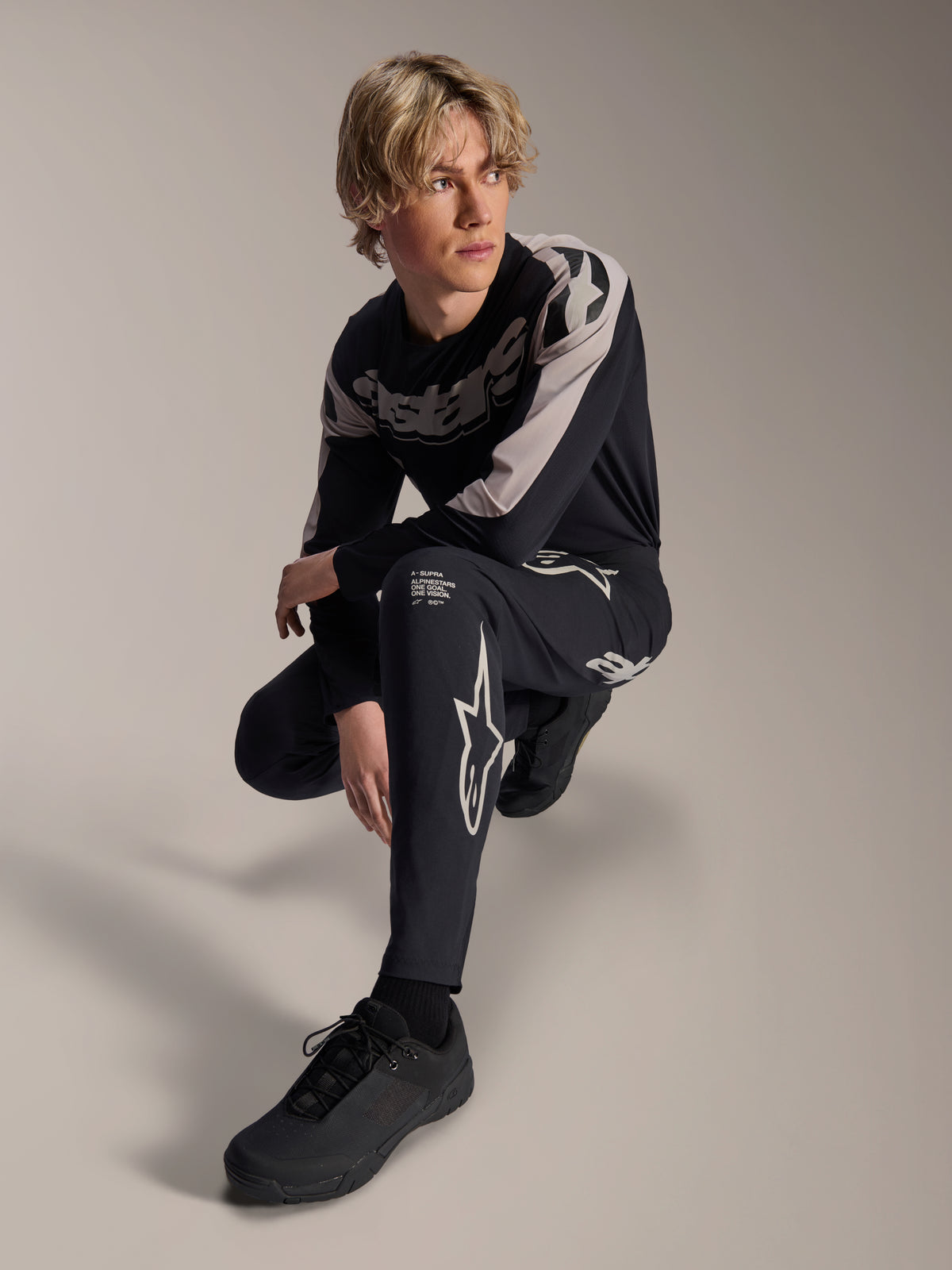 Alpinestars A-Supra Melt bukser og langærmet jersey, sort og grå, med markant hvid branding og stjernelogoer, båret af en mandlig model i en hugsiddende positur, koordineret MTB-racerudstyr