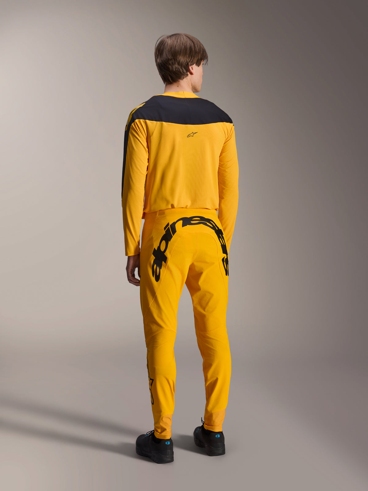 Alpinestars Limited Edition A-Supra Melt Gold-udstyrssæt, MTB-trøje og bukser, mørkegul og sort, set bagfra med stort Alpinestars-navnetræk på bagstykket og sort bærestykke på trøjen, performance mountainbike-tøj
