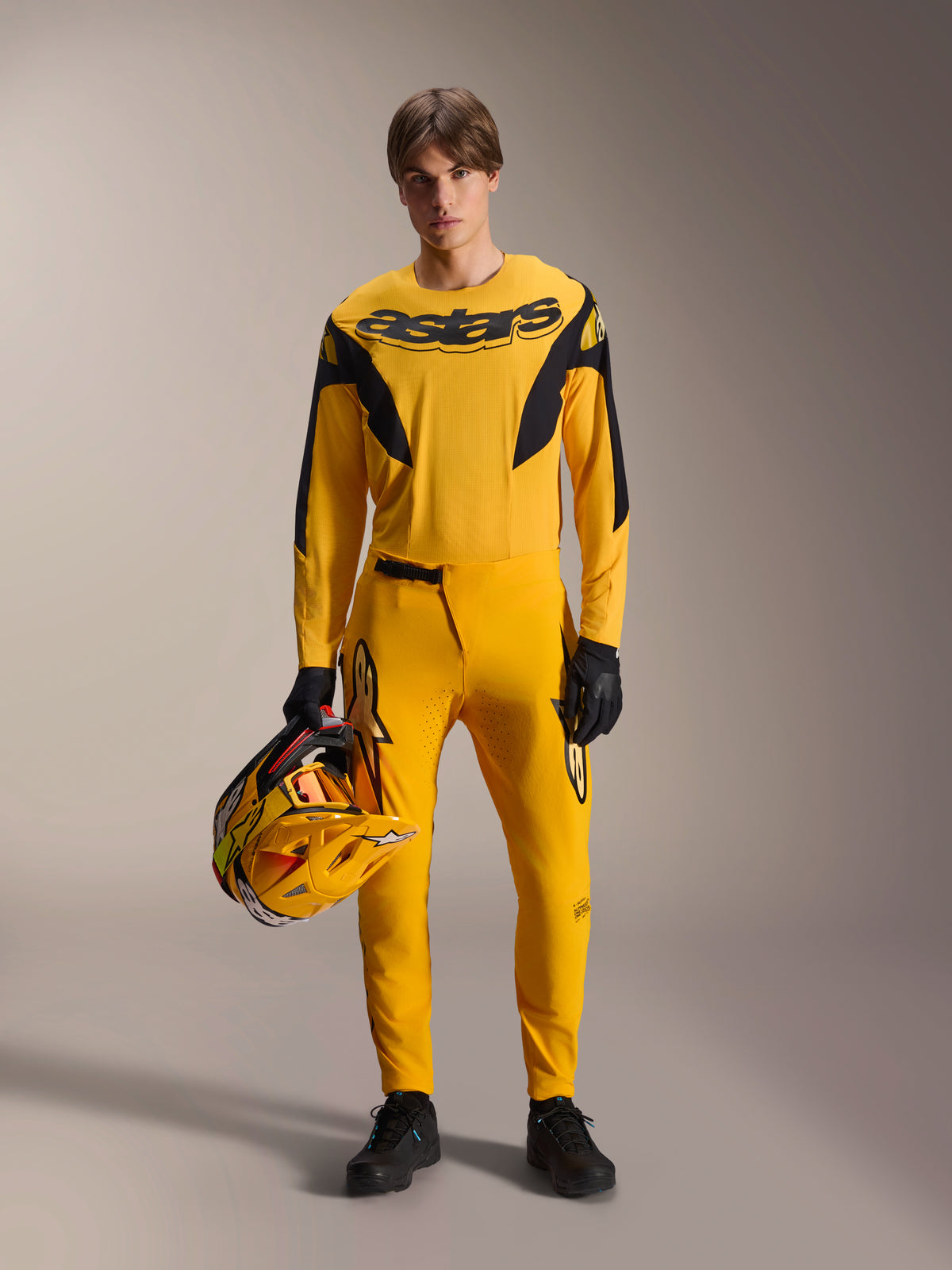 Model iført Alpinestars Limited Edition A-Supra Melt Gold MTB-sæt, gul langærmet jersey med sort 'astars'-logo, matchende gule bukser med sorte detaljer, holder en gul og sort racerhjelm, sorte handsker og sko, performance-udstyr til mountainbiking