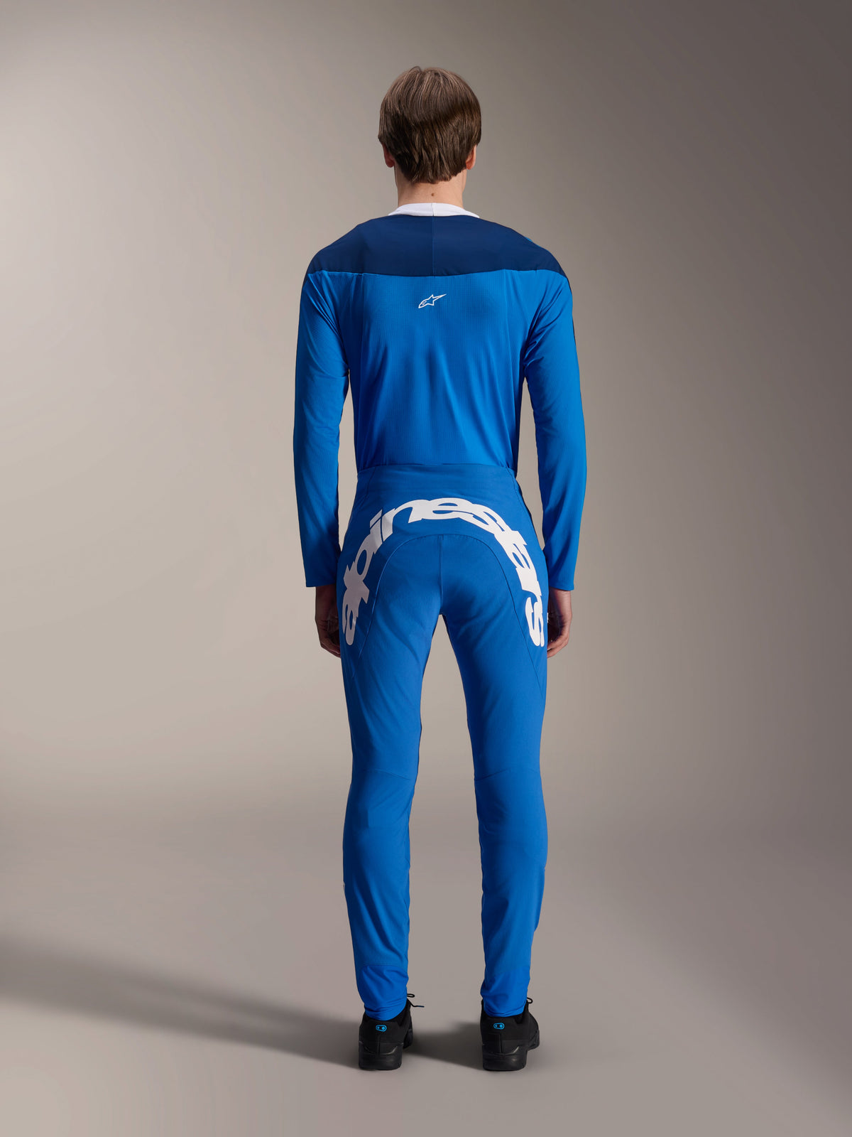 Mandlig model set bagfra iført A-Supra Melt Jersey - Long Sleeve i blå, mørkeblåt skulderpanel med et lille hvidt stjernelogo, matchende blå MTB-bukser med stort hvidt Alpinestars-branding over bagdelen, koordineret performance-cykeloutfit til mountainbike.