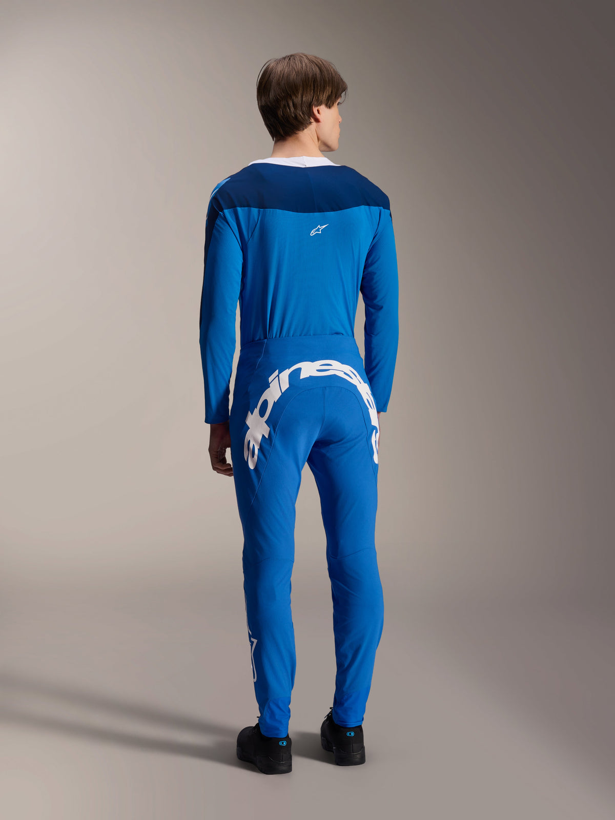 Mandlig model set bagfra iført Alpinestars A-Supra Melt langærmet trøje i blå, kombineret med matchende blå præstationsbukser med et stort hvidt Alpinestars-logo hen over ryggen, komplet MTB-racerkit