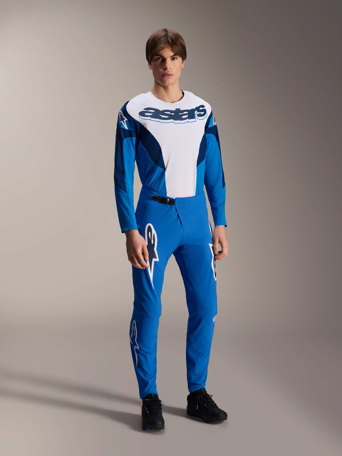 Mandlig model iført Alpinestars A-Supra Melt langærmet trøje i blå og hvid, med et markant 'astars'-logo på brystet, matchende blå mountainbike-bukser og sort fodtøj, ydelsesorienteret MTB-racerbeklædning.