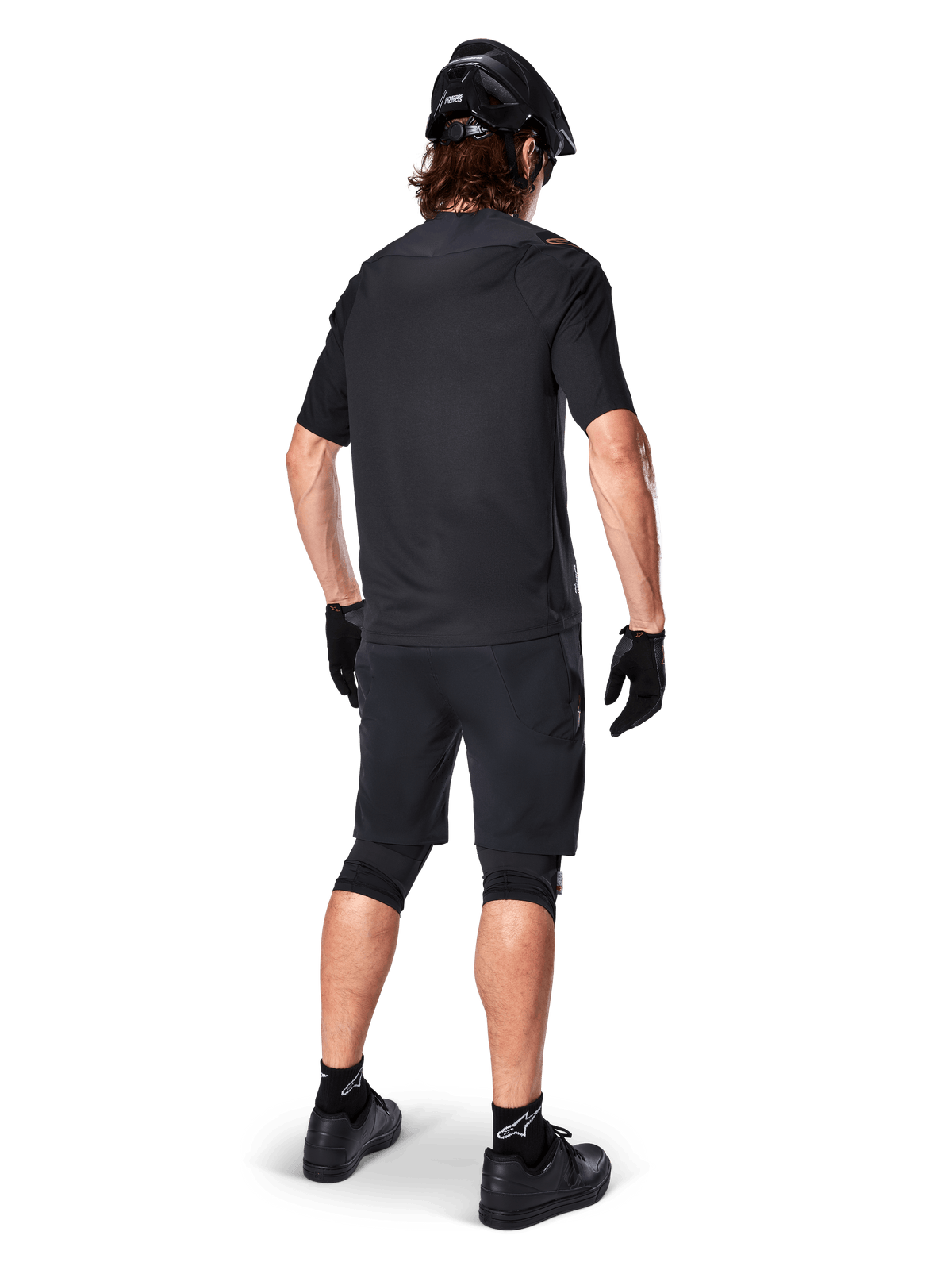 Mandlig mountainbikerytter set bagfra iført Alpinestars A-Aria Polartec Switch Jersey i sort, kortærmet MTB-trøje, kombineret med sorte mountainbike-shorts, sort hjelm og handsker, koordineret performance-outfit til terrænkørsel på stier.