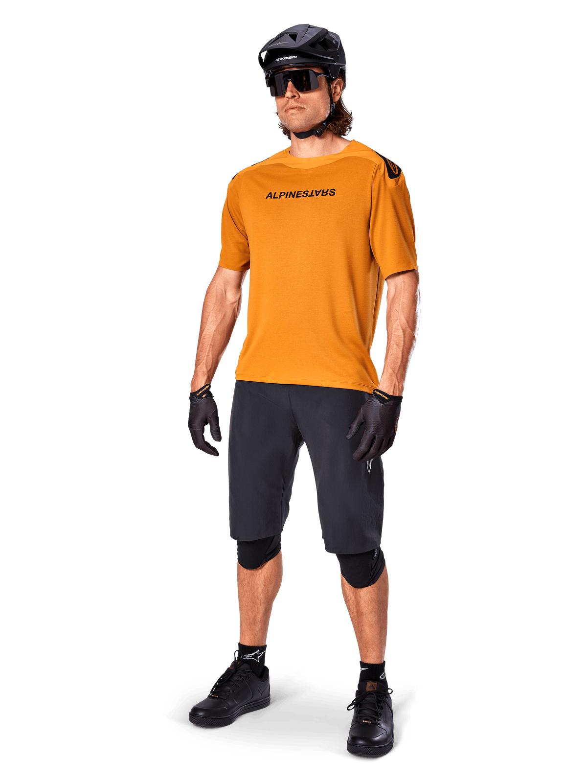 Male rider wearing the Alpinestars A-Aria Polartec Switch Jersey in Dark Gold, short-sleeve MTB jersey, paired with black cycling shorts, knee guards, black helmet, and gloves, professional mountain bike outfit for trail riding