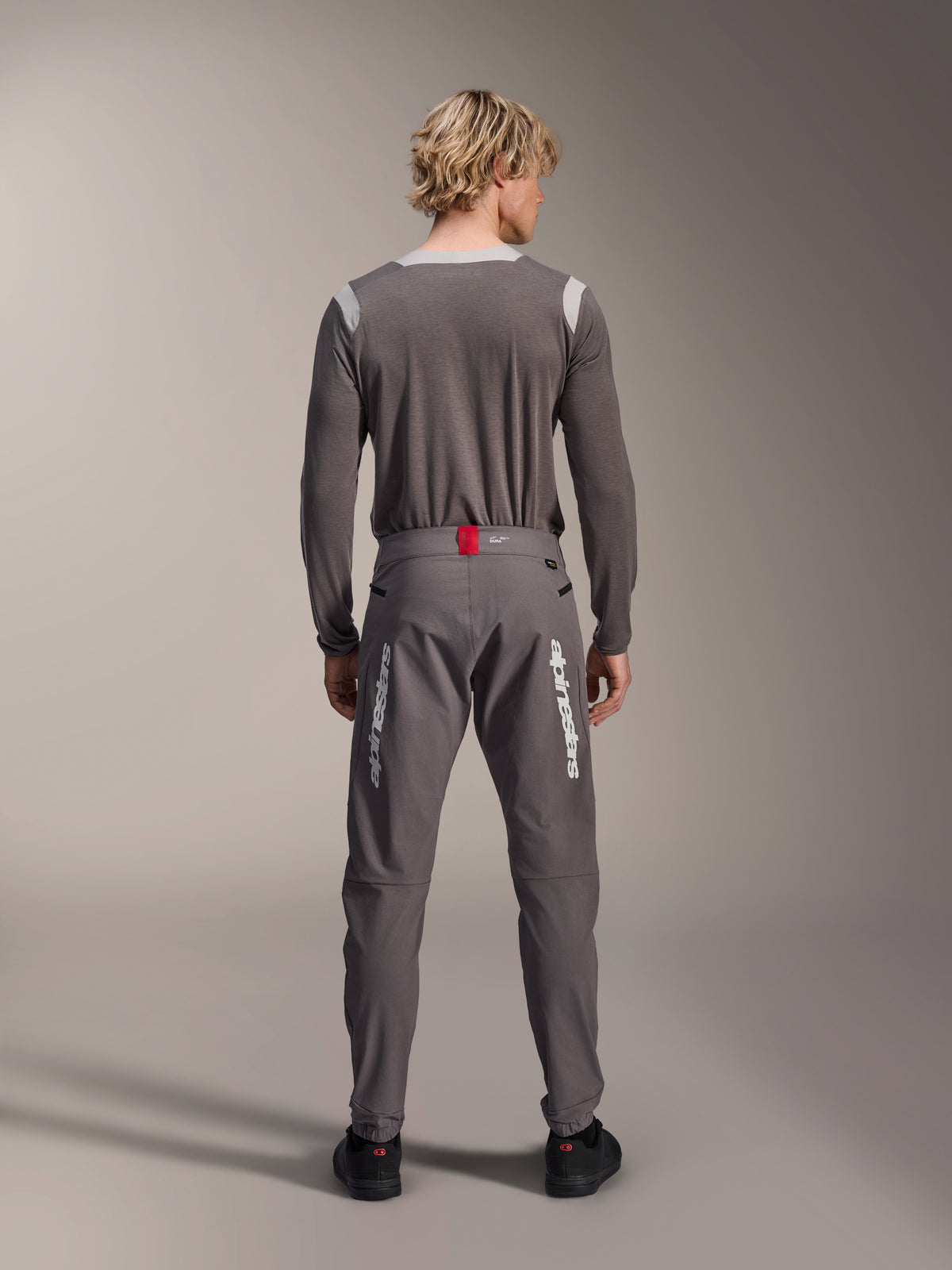 Model set bagfra iført Alpinestars A-Dura Elite bukser i mørkegrå og en matchende grå langærmet trøje, med hvide Alpinestars-logoer på benene, en rød detalje på linningen og sorte sko, professionelt MTB-udstyr.