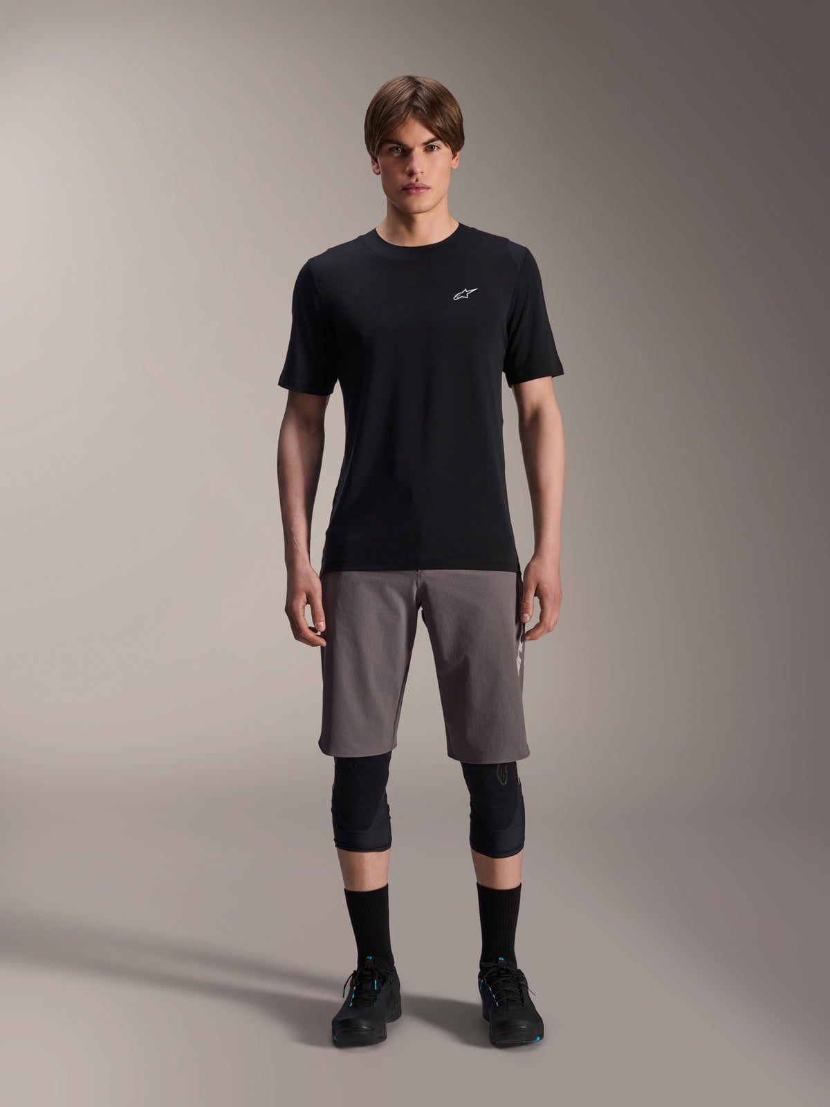 Alpinestars A-Dura Dri Wool kortærmet jersey, sort, båret af en model med grå mountainbike-shorts, sorte knæbeskyttere og sorte cykelsko, minimalistisk design med et lille hvidt Astars-logo på brystet, funktionelt performance MTB-tøj til trailkørsel.